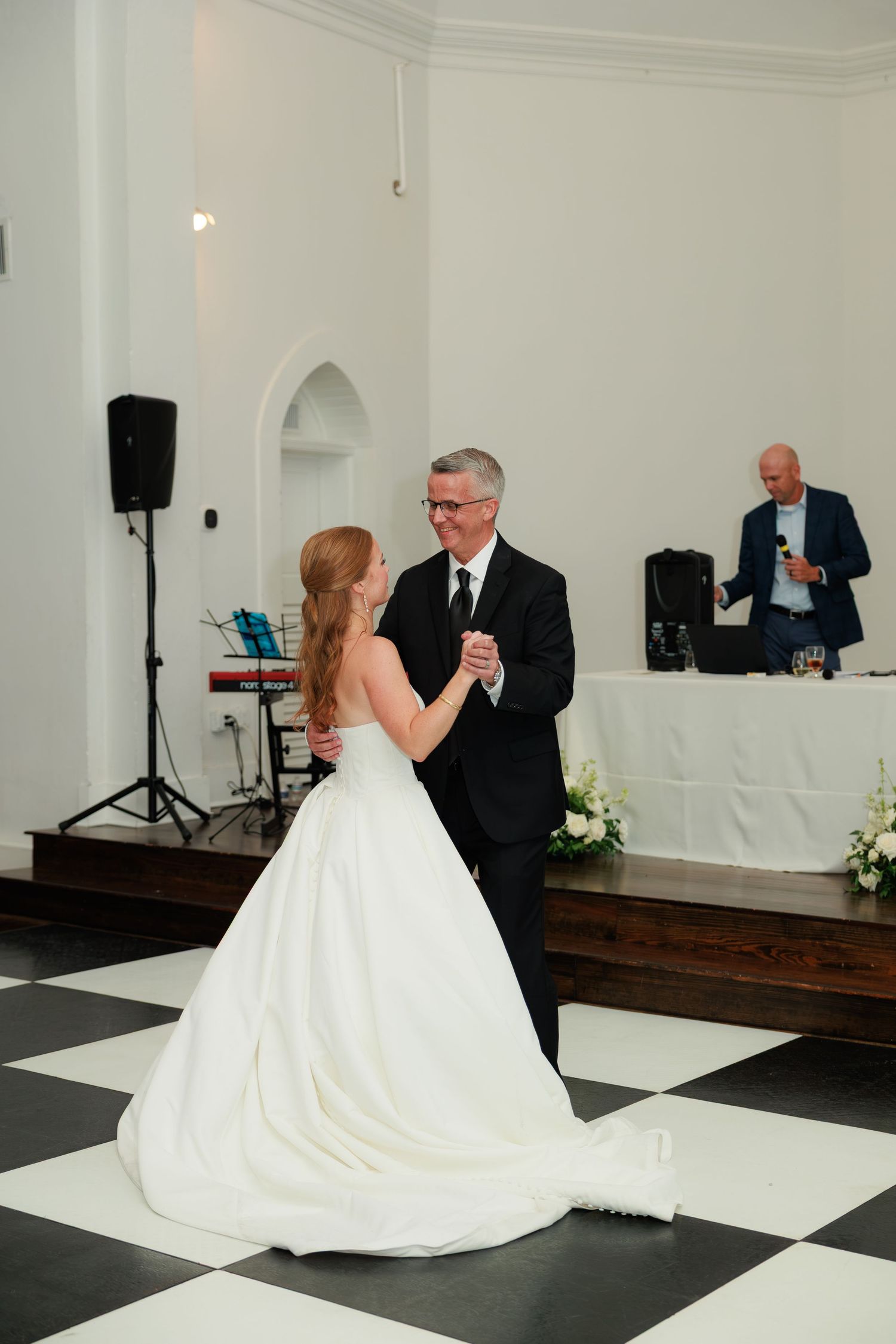 Bride in white gown shares father-daughter dance on checkered floor at elegant church wedding reception.