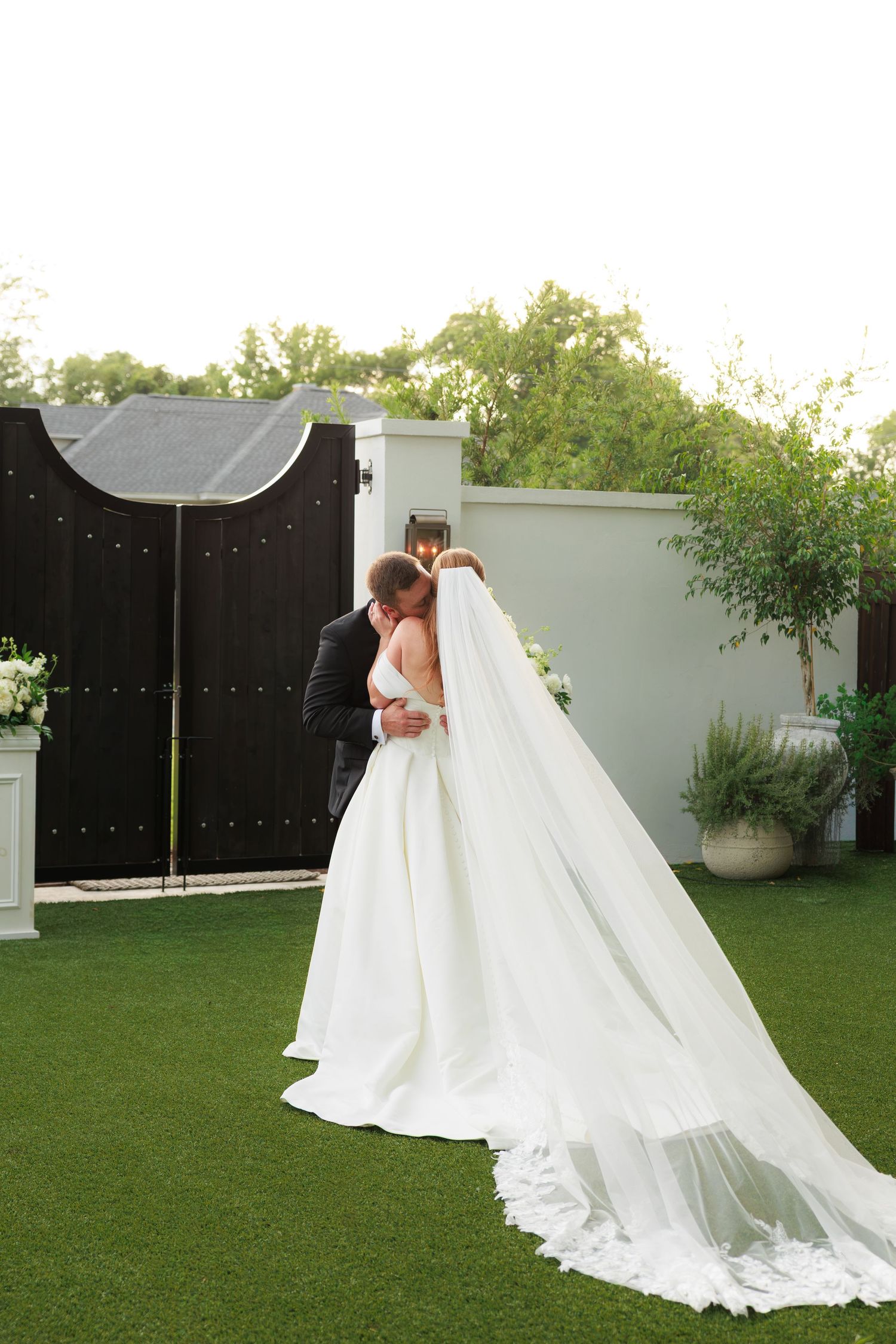 Bride and groom share first kiss in lush garden ceremony venue with elegant black gates and string lights.