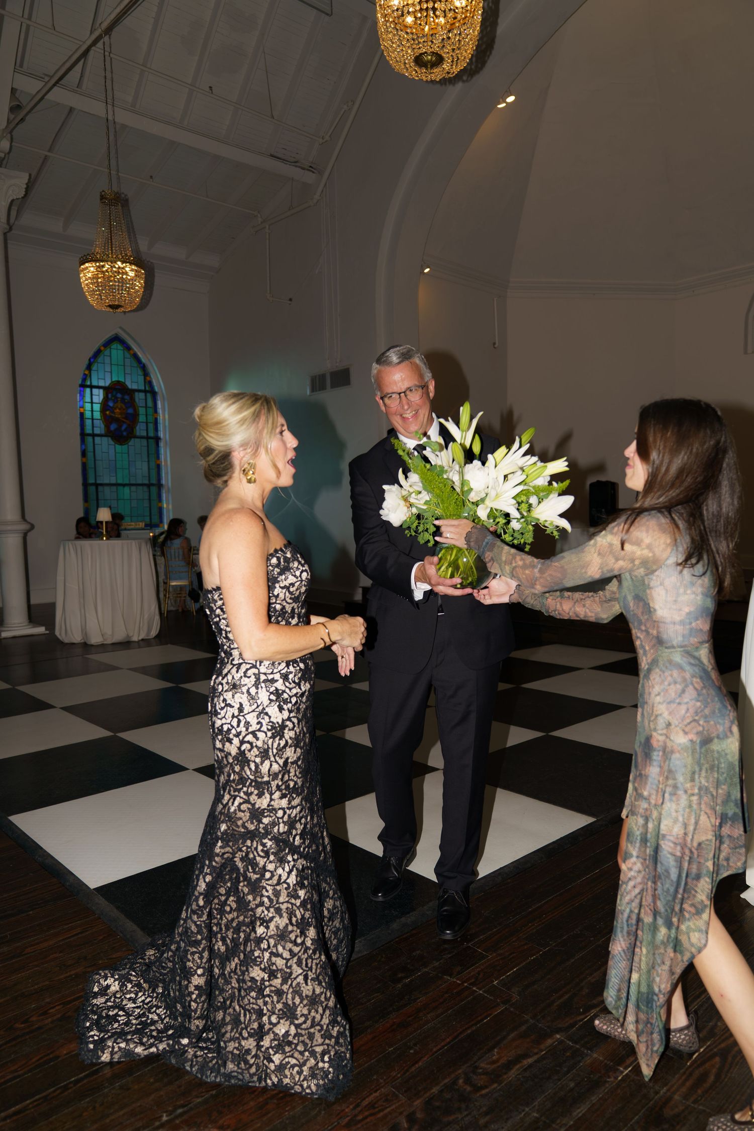 Elegant guests in formal attire exchange a bouquet of white lilies on a checkered dance floor inside a decorated venue.
