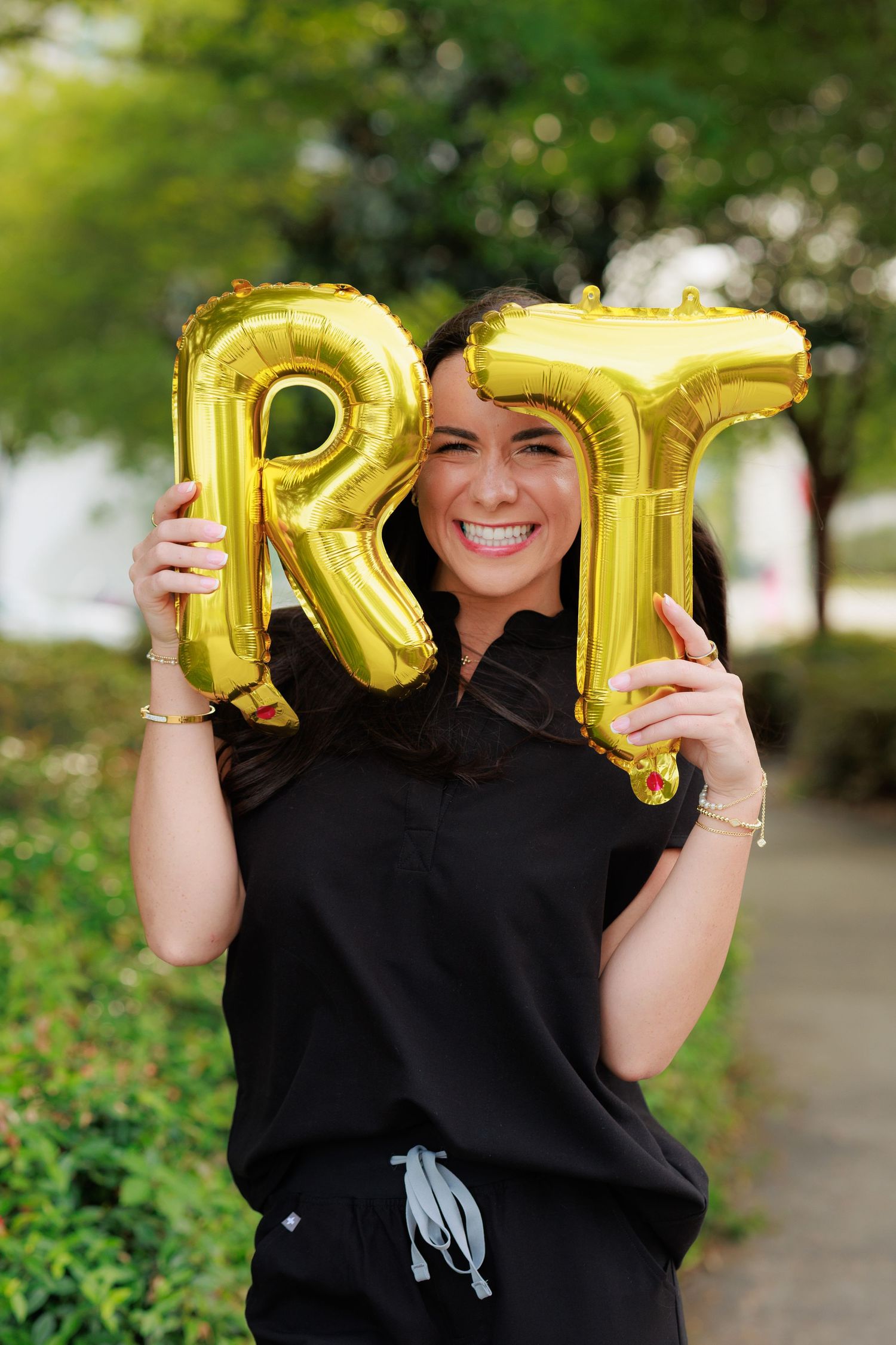 Smiling woman in black scrubs holding gold RT letter balloons outdoors, celebrating respiratory therapist graduation.