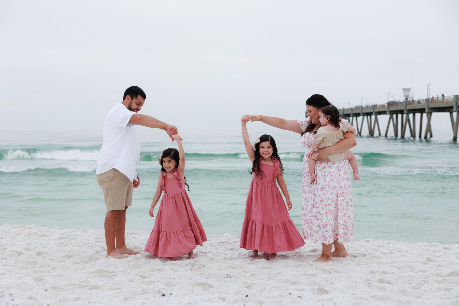 Family of four posing on white sand beach near pier, parents twirling daughters in matching pink dresses by turquoise ocean.