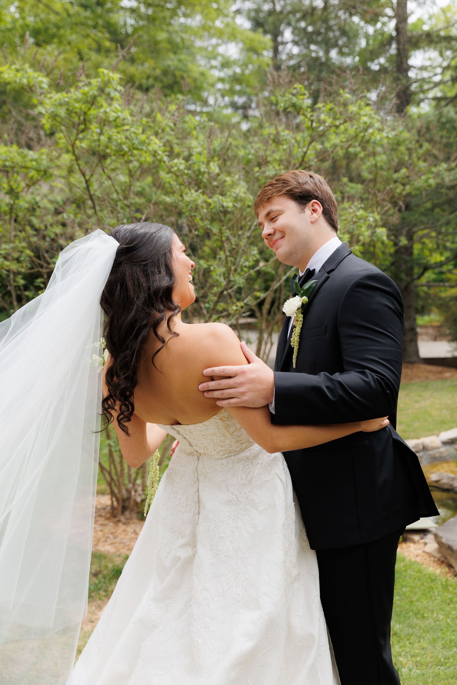 Smiling groom in black tuxedo embraces bride in white lace gown during outdoor garden first look.