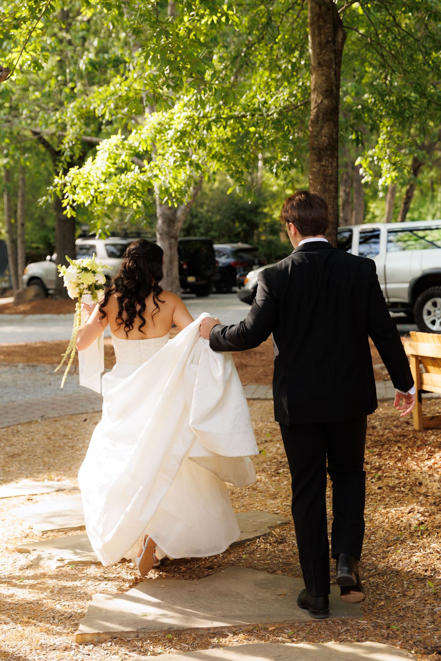 Bride in white gown holding bouquet walks with groom in black suit through tree-lined path at outdoor wedding.