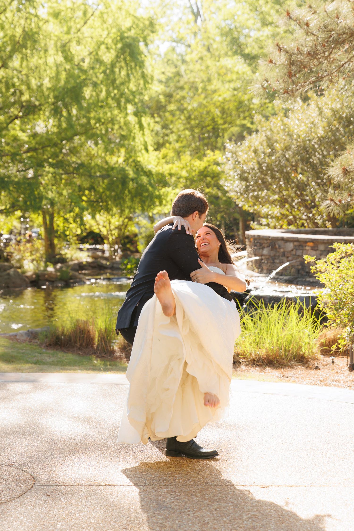 Groom lifts smiling bride in white wedding dress during a playful outdoor moment by a garden pond.