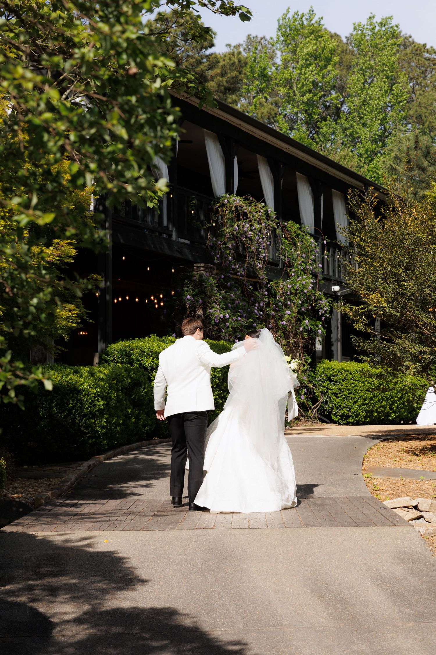 Newlywed couple walking hand in hand toward elegant ivy-covered black wedding venue surrounded by trees.