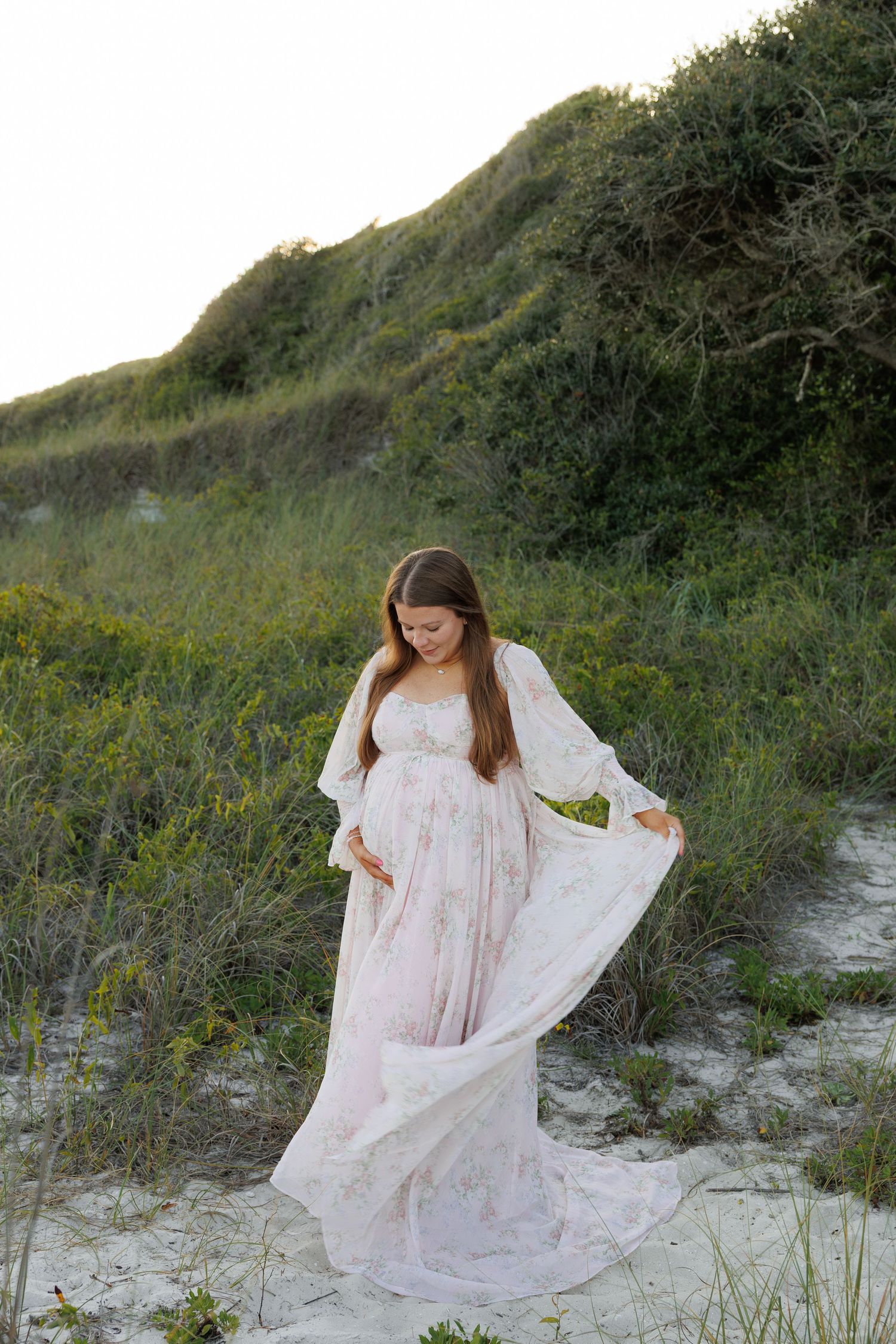 Pregnant woman in floral gown holding belly on sandy beach dunes with lush greenery backdrop.