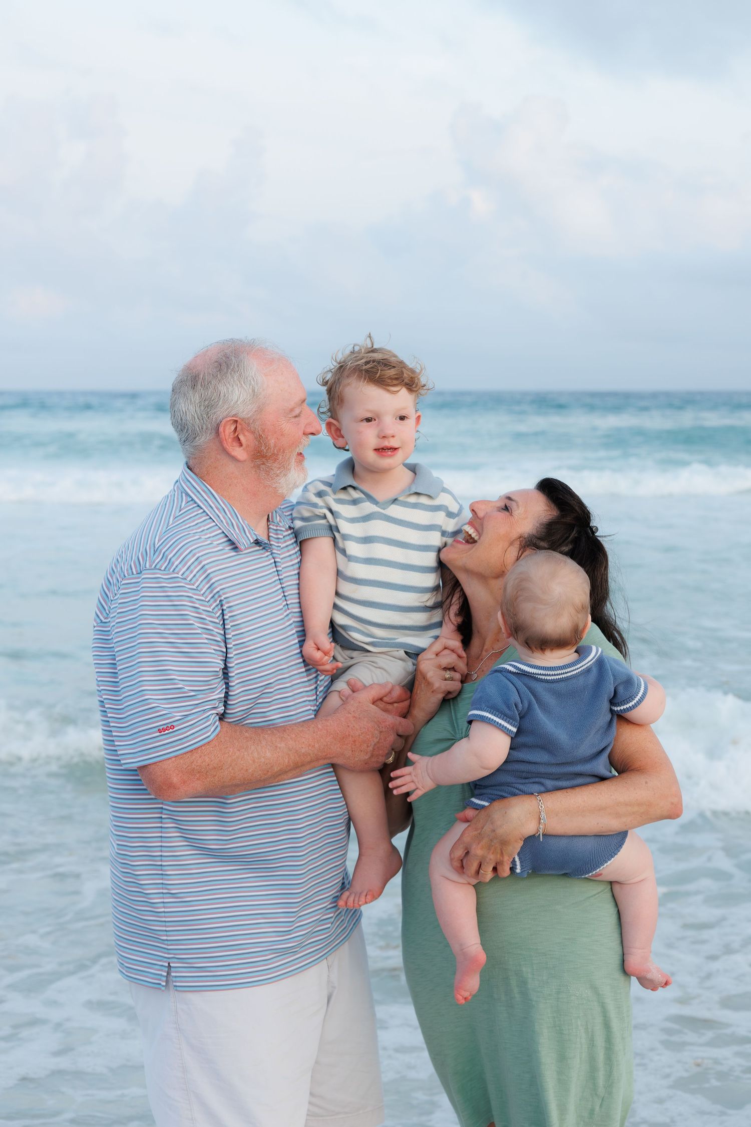 Grandparents laughing with two toddler grandchildren on beach with turquoise water.