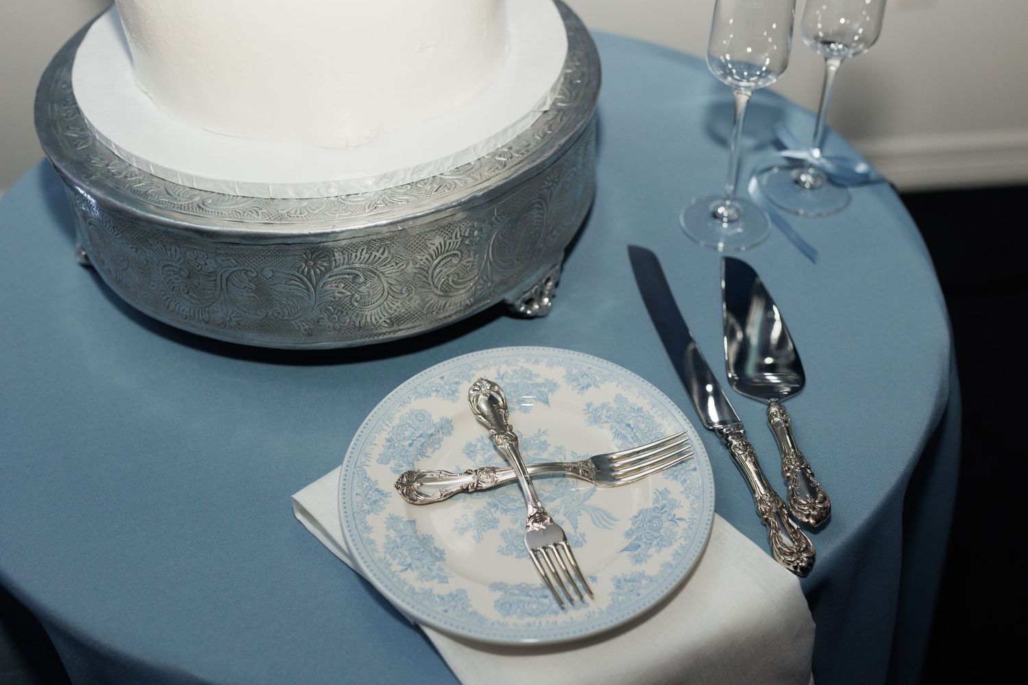 An elegant place setting with silver utensils and a decorative blue and white plate beside a round silver cake stand.