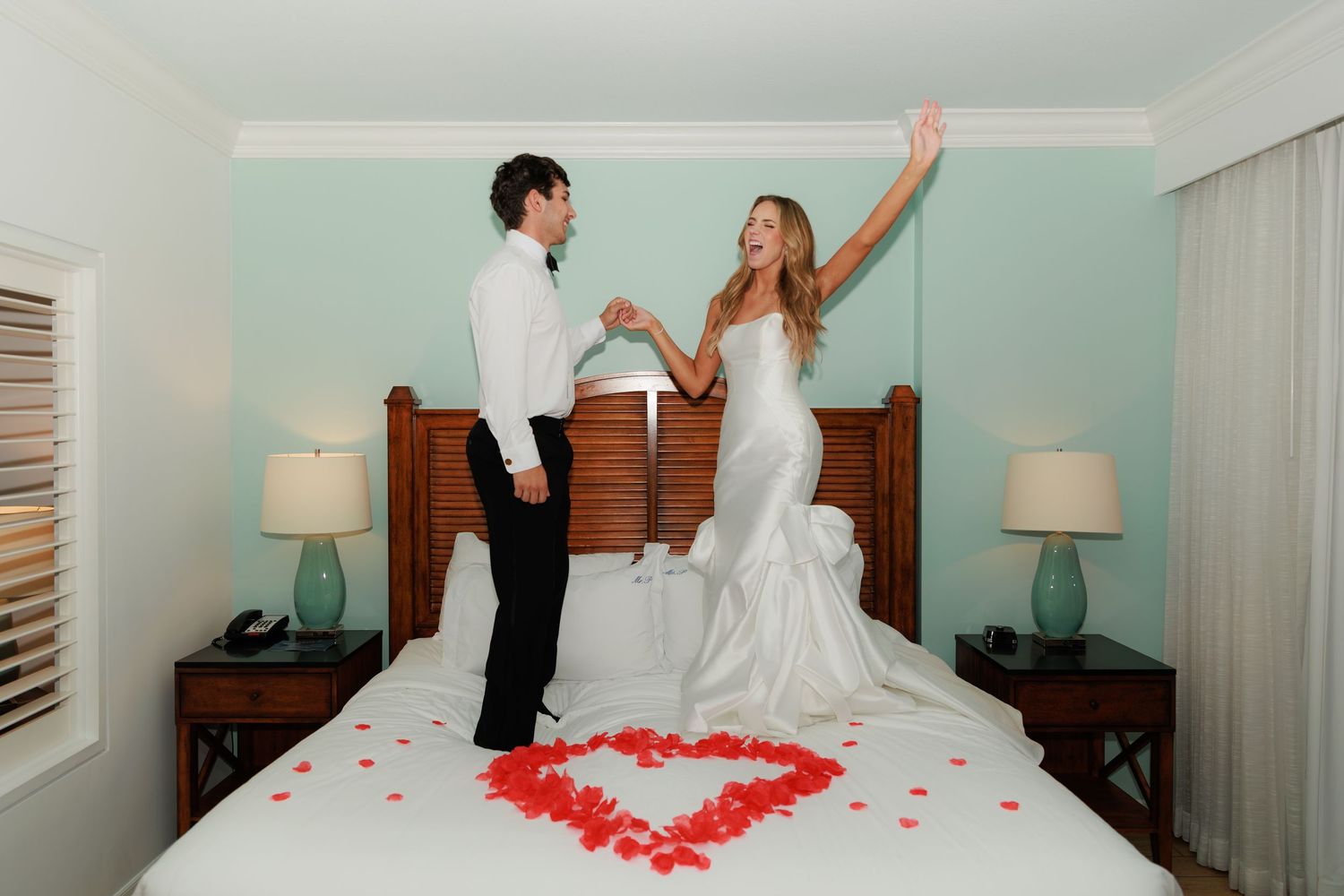 A couple celebrates on a bed decorated with rose petals arranged in a heart shape.