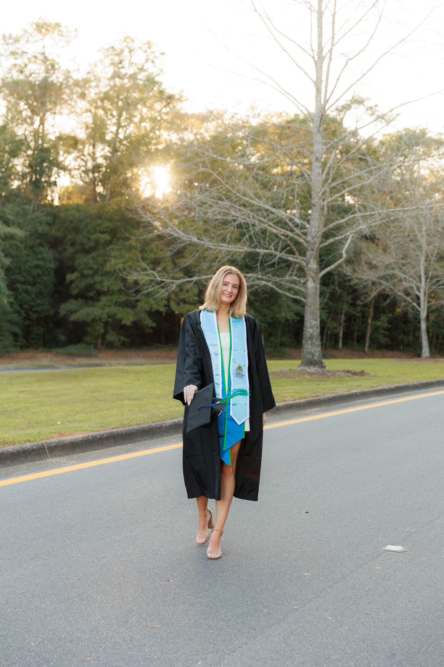 Graduate in black cap and gown walking on a road at sunset with trees in the background.