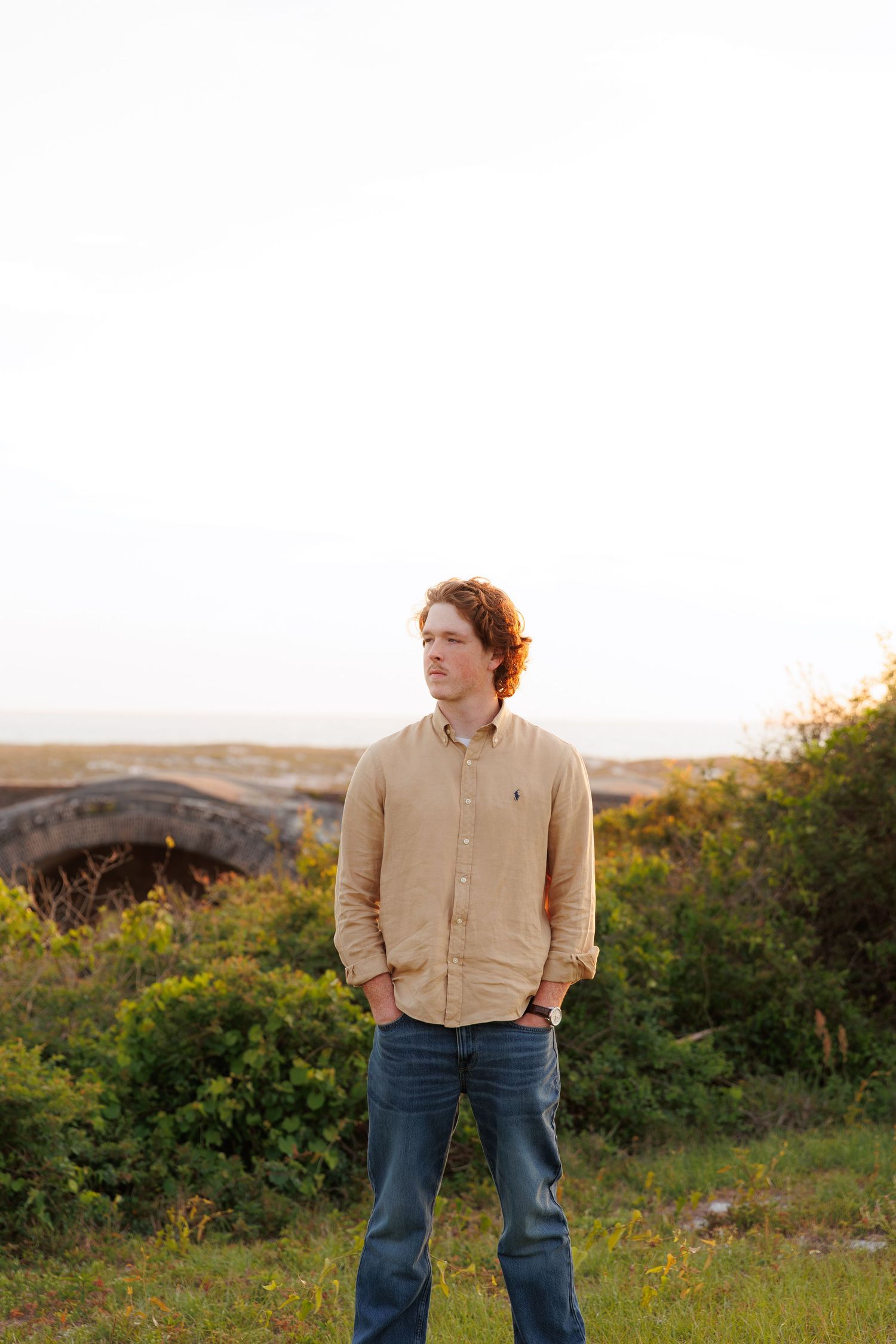 Young man with curly red hair in beige shirt and jeans stands outdoors near coastal greenery at golden hour.
