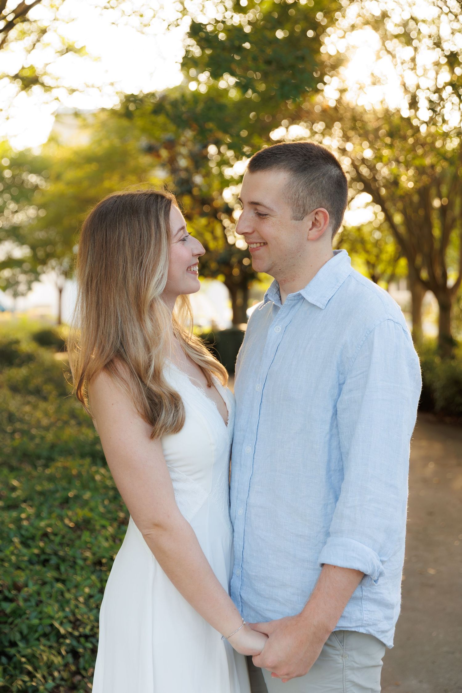 Smiling couple holding hands in a sun-dappled park, woman in white dress and man in light blue shirt.