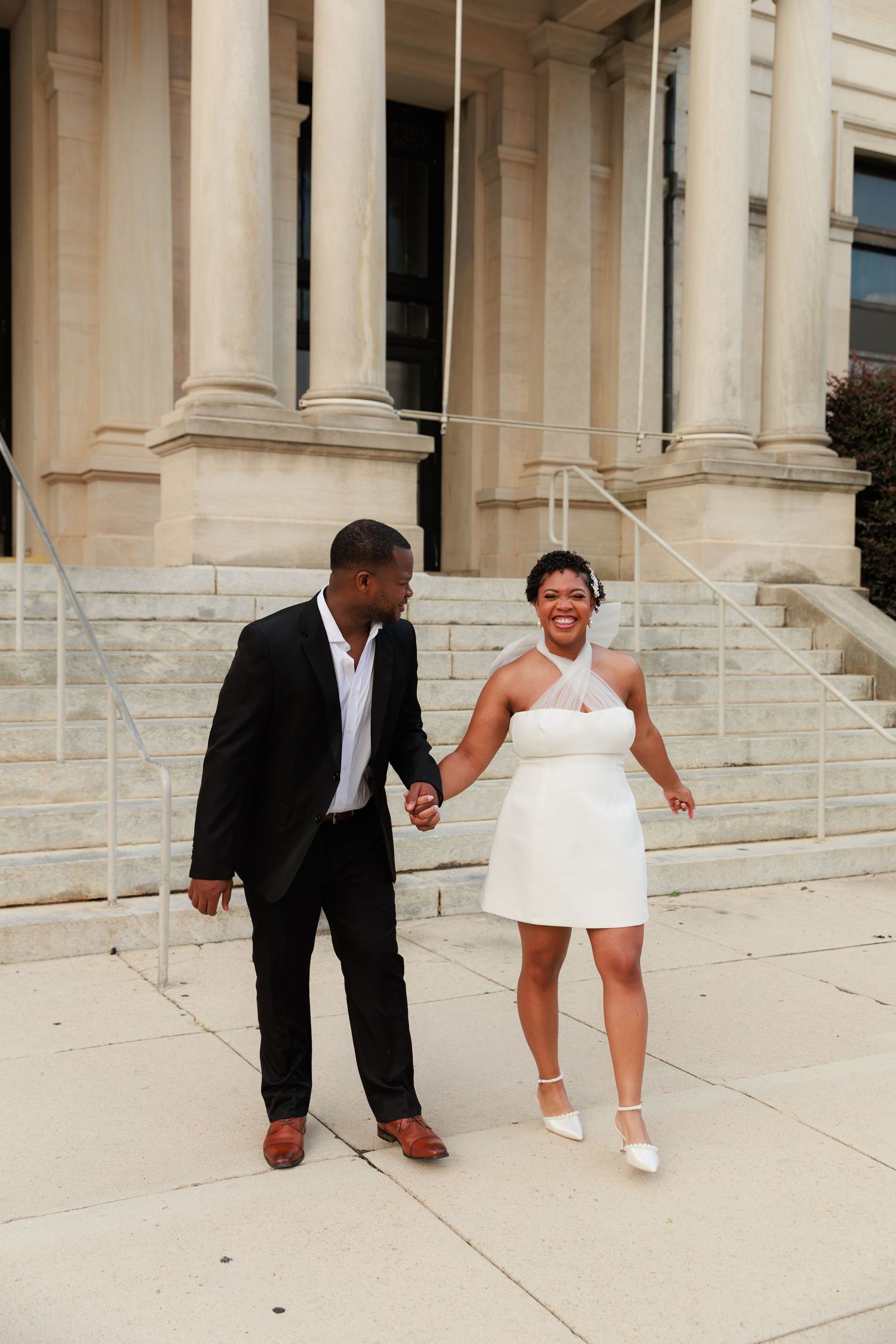 A joyful Black couple holds hands outside a neoclassical building, bride in a short white halter dress and groom in a dark suit.