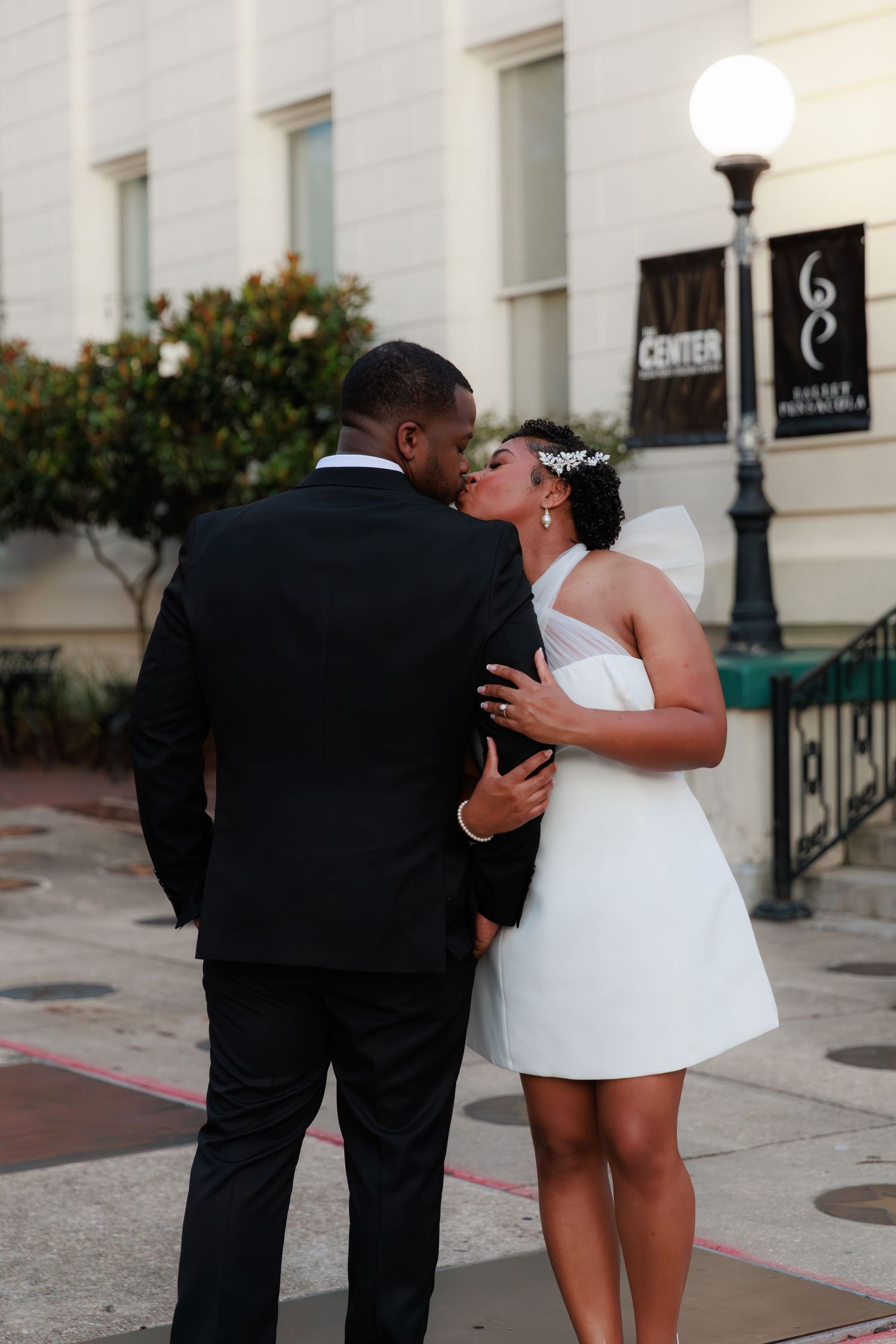 A newlywed couple kiss tenderly outside The Center building, bride in a short white dress with veil and groom in a black suit.