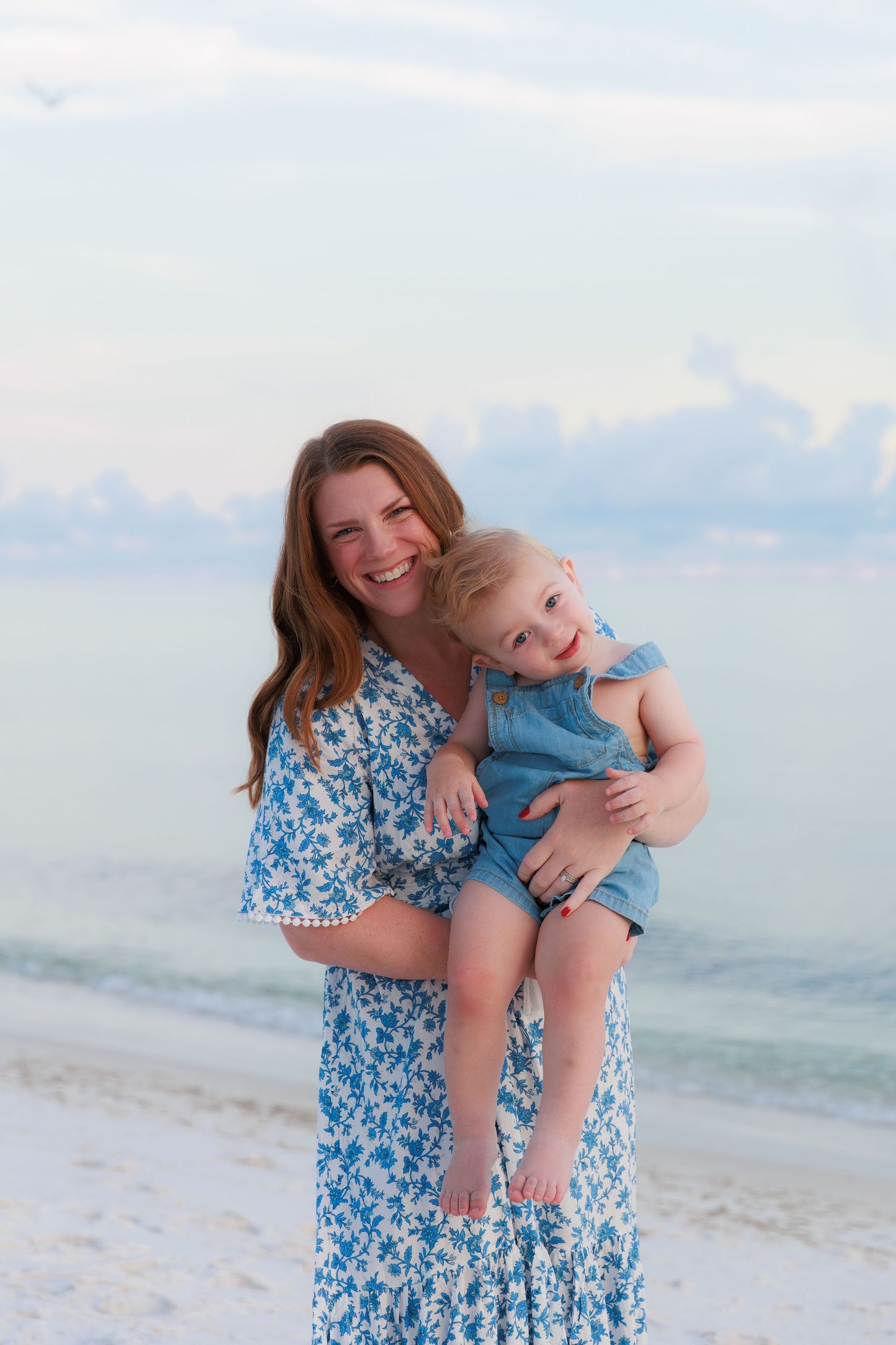 A smiling woman in a blue floral maxi dress holds a laughing toddler on a white sand beach at dusk.