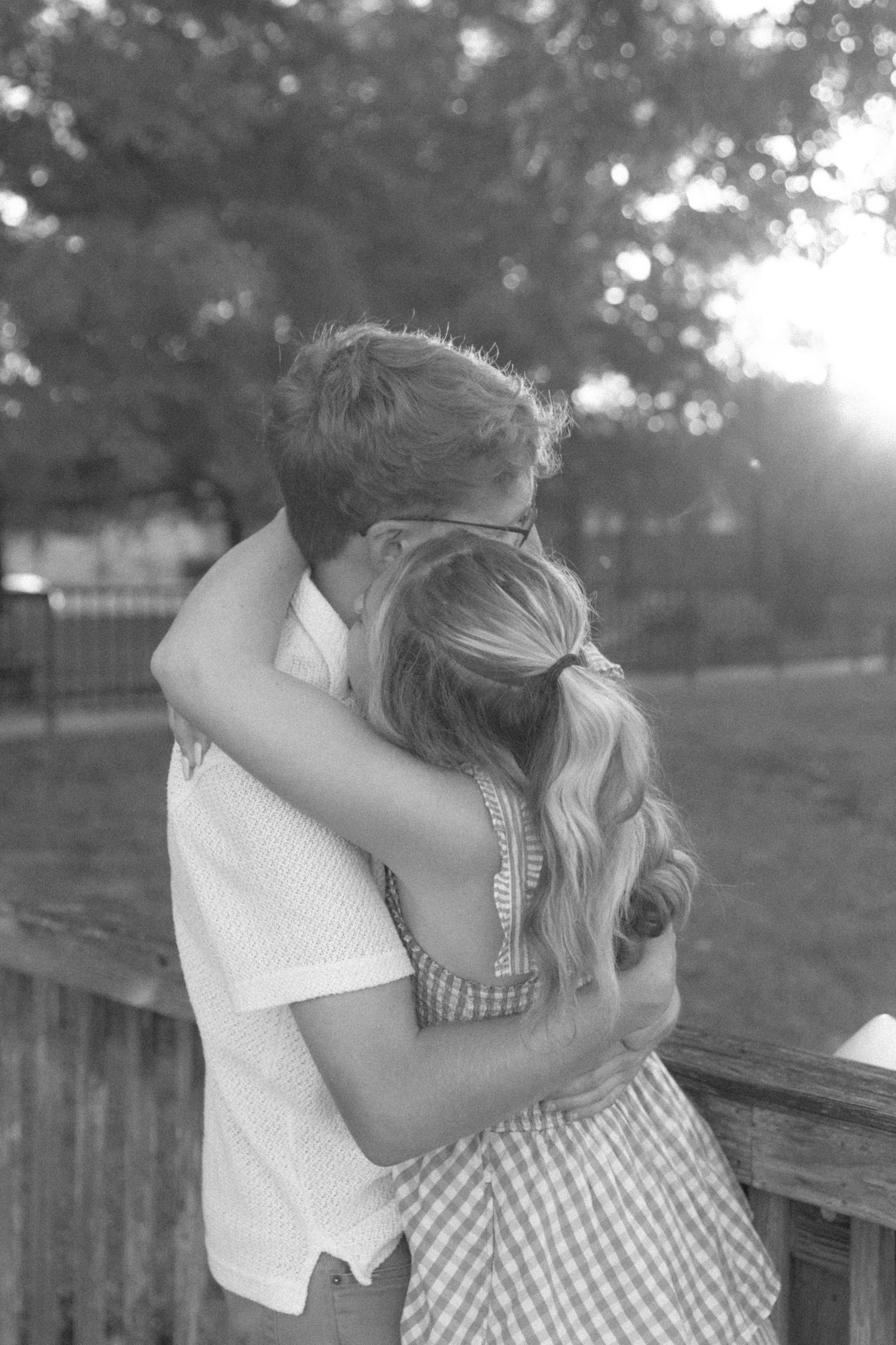Black and white photo of a couple embracing and kissing on a wooden bridge surrounded by trees.