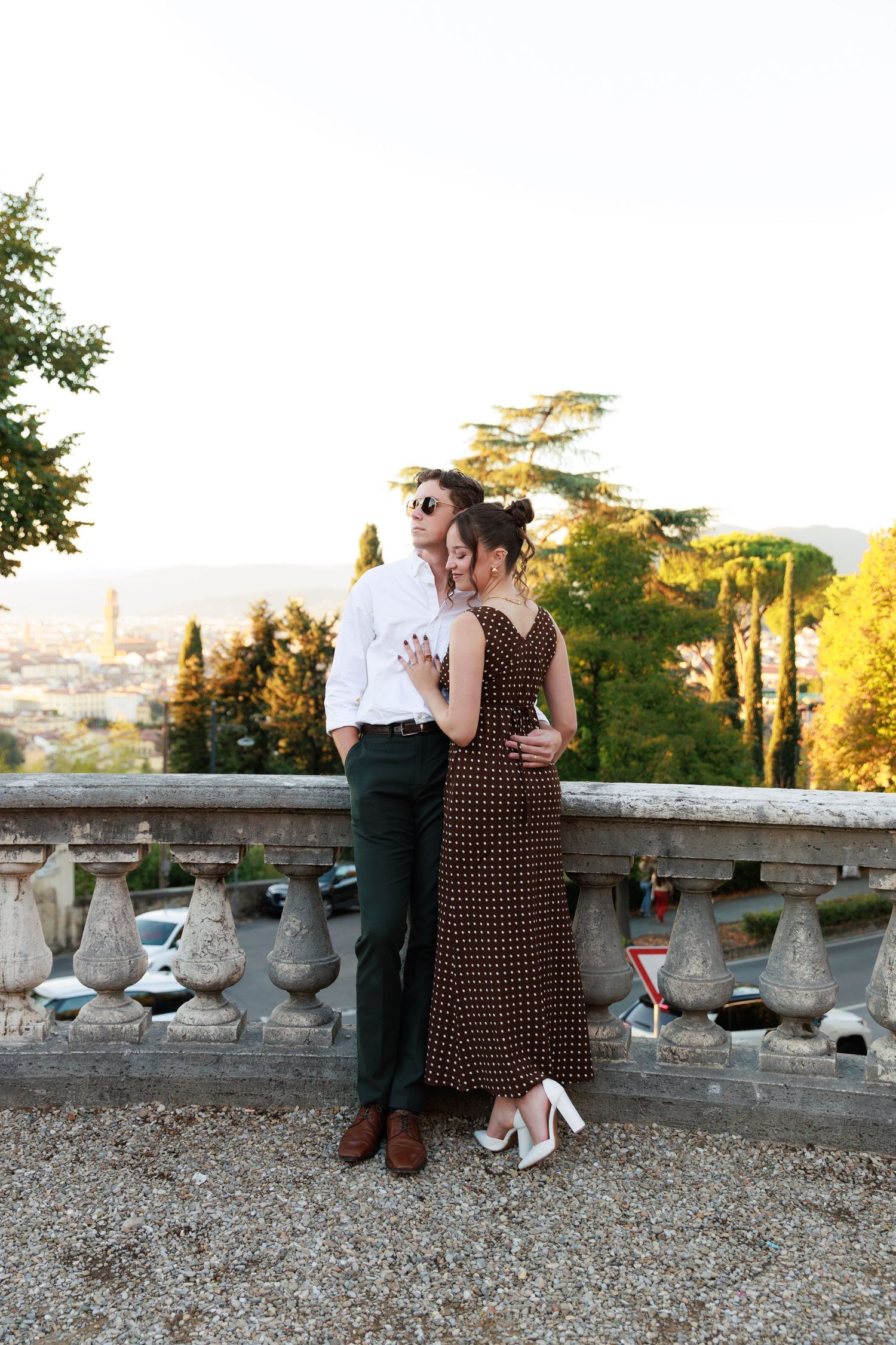 Elegant couple embracing on a terrace overlooking Florence cityscape at sunset, woman in polka dot maxi dress.