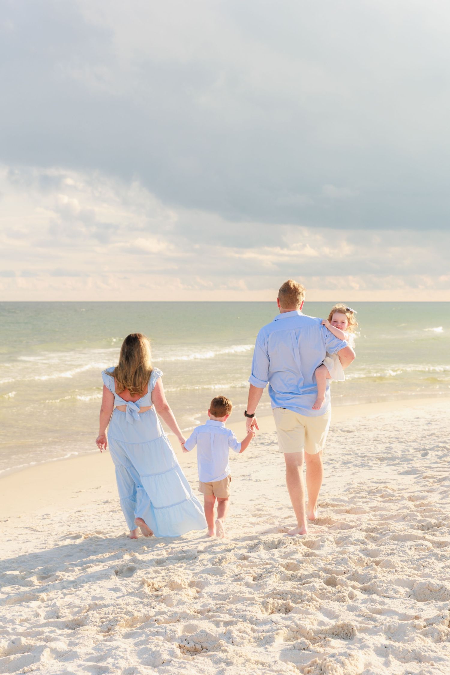 Family of four walking hand-in-hand on a sandy beach toward ocean waves, dressed in coordinating blue and white outfits.