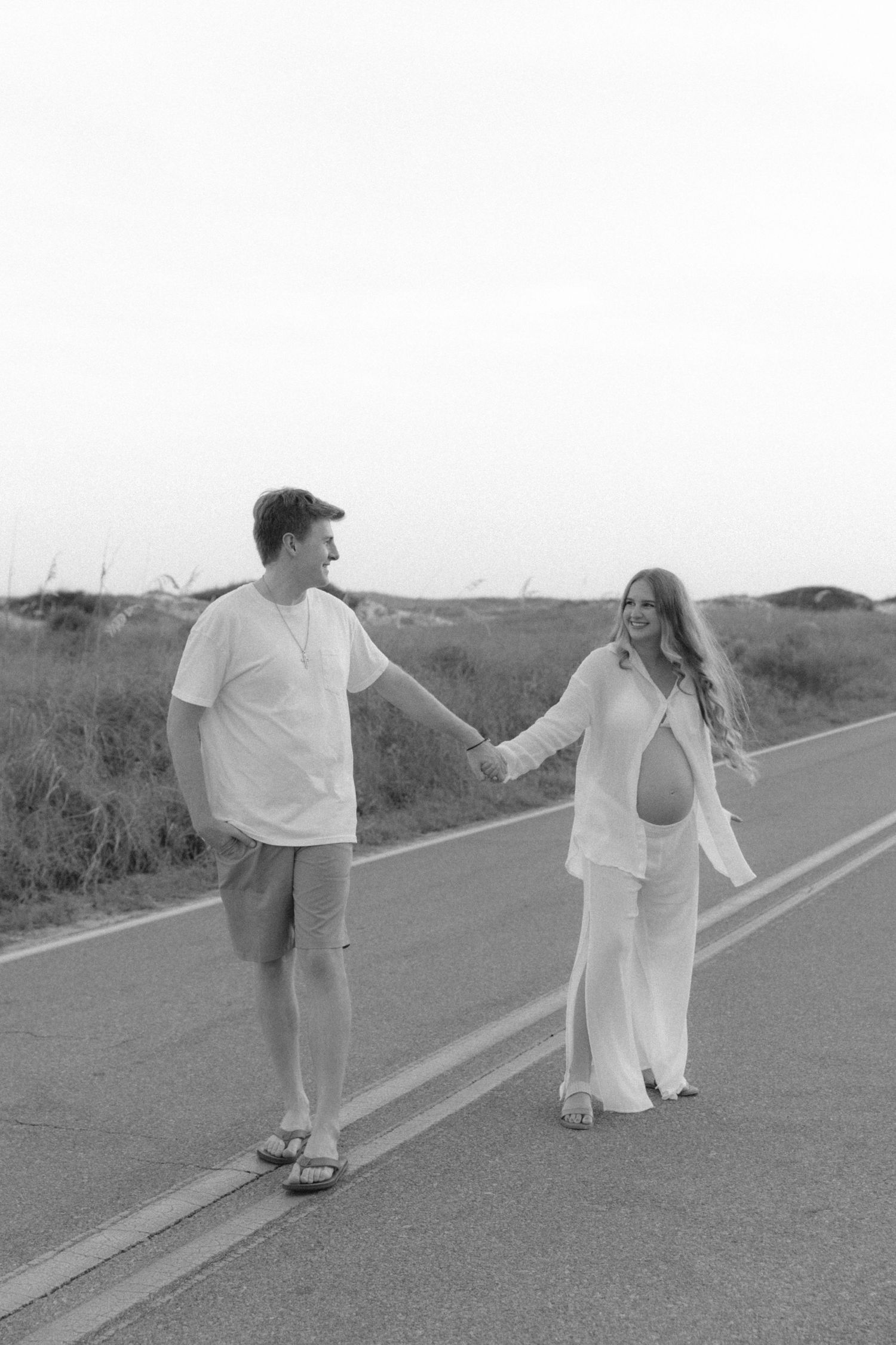 A couple walks together on a coastal road at sunset, with the pregnant woman wearing a flowing white dress.