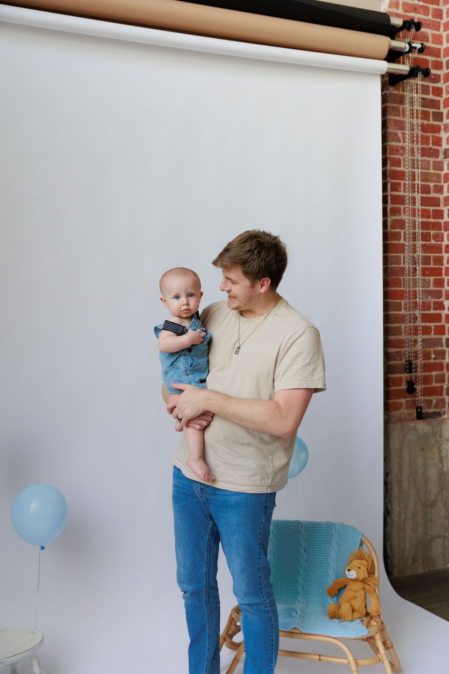 A parent holds a baby in casual attire while a blue balloon and stuffed animal decorate the scene against a white wall.