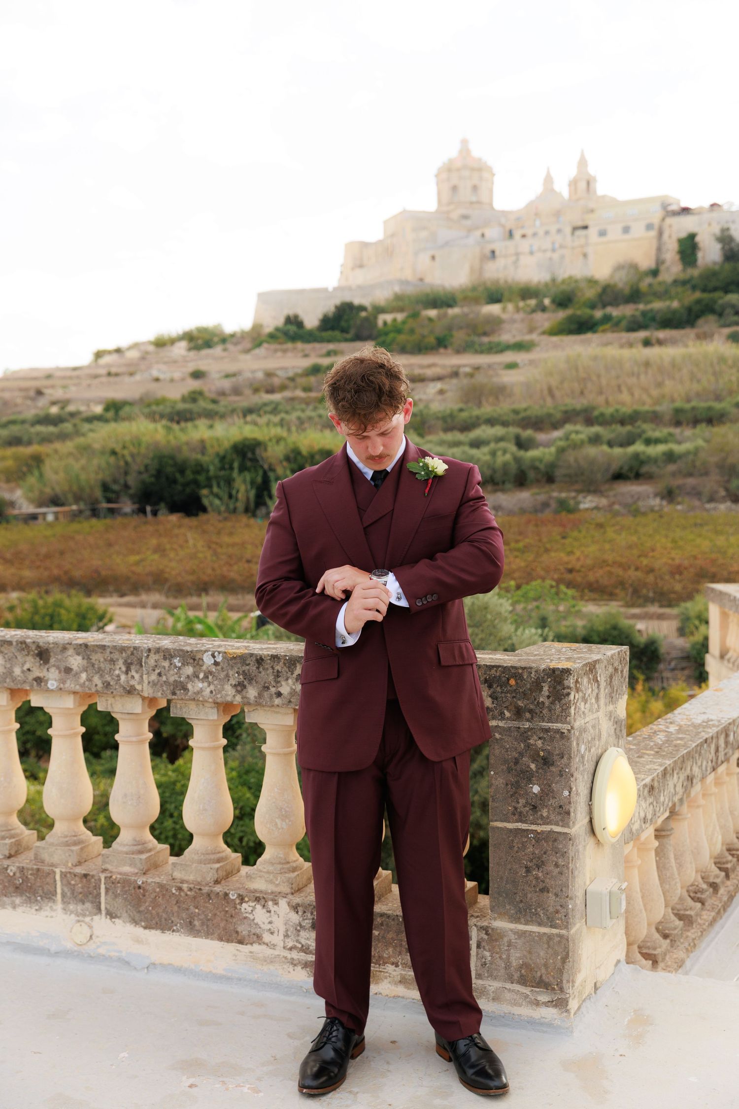 A person in a burgundy suit checks their watch while standing on a stone balcony with a scenic hillside view.