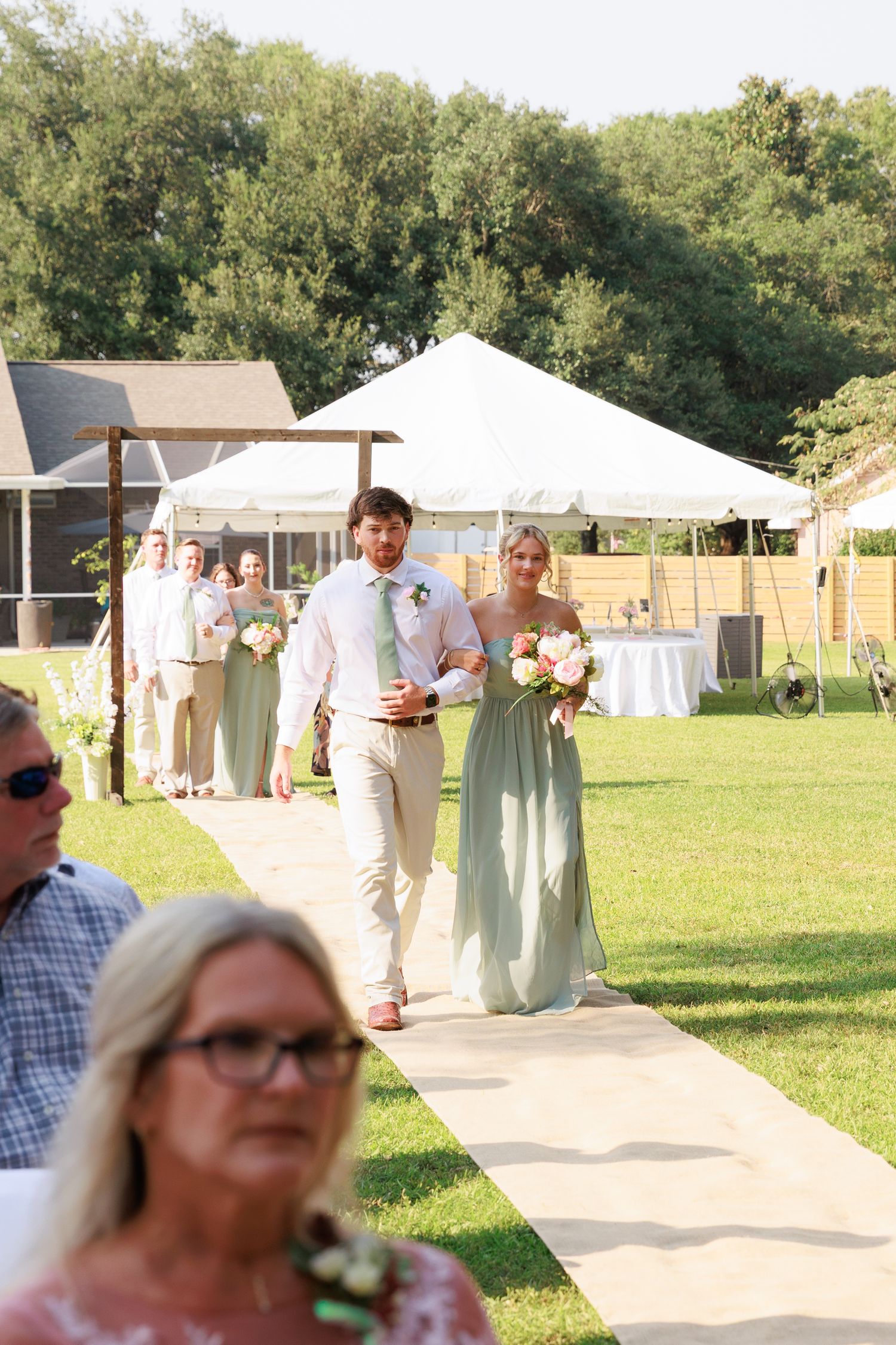 Outdoor wedding reception setup with white tents and umbrellas scattered across a grassy field on a sunny day.