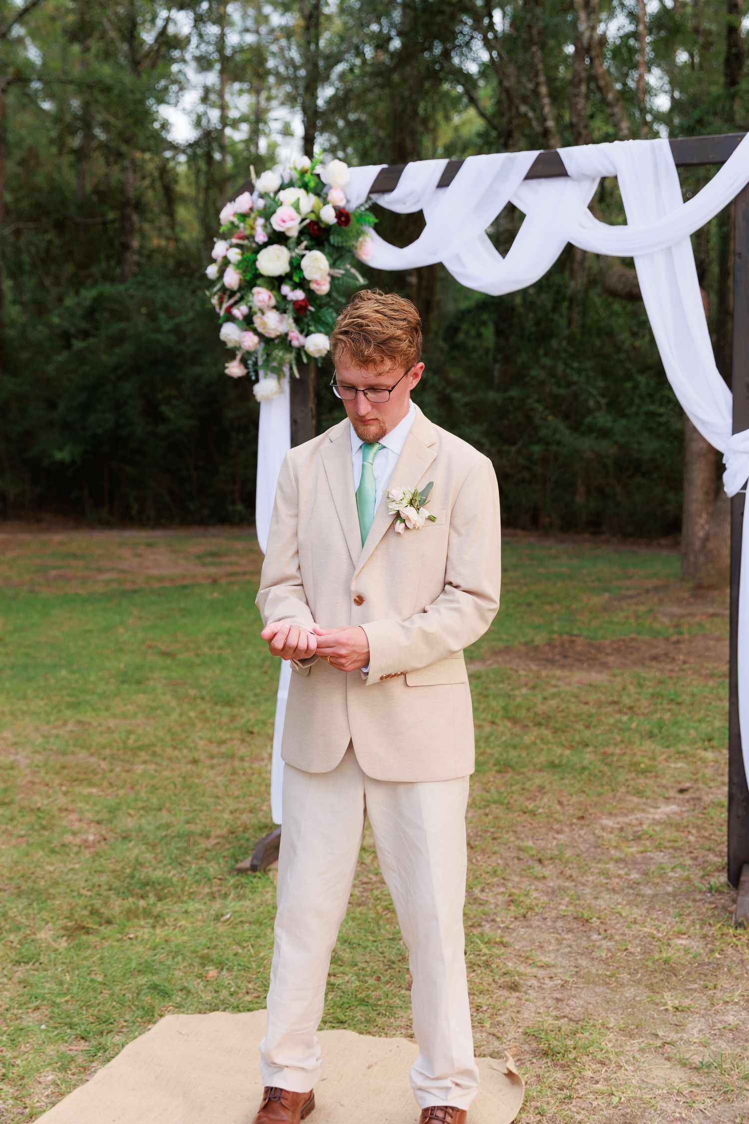 Groom in light beige suit checking phone under white wedding arch in outdoor garden setting.