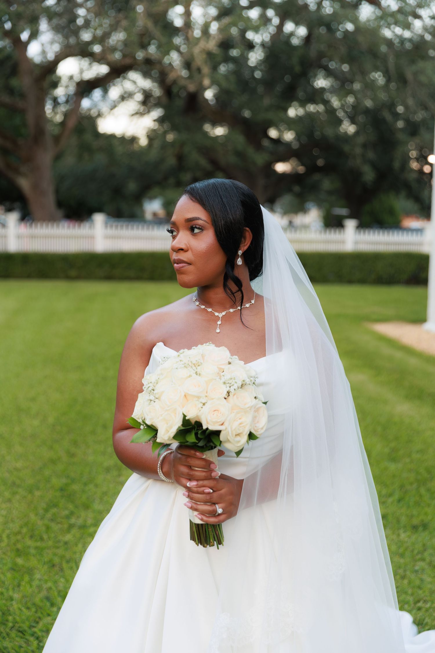 Beautiful bride in white wedding gown holds bouquet of white roses while posing in outdoor garden setting.