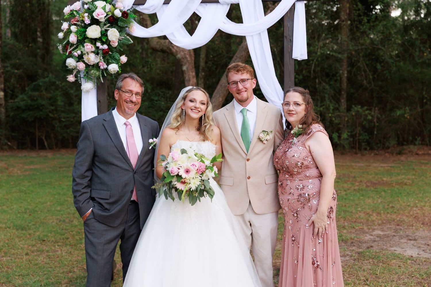 Wedding party poses outdoors in front of white monogram backdrop with pink floral arrangements.