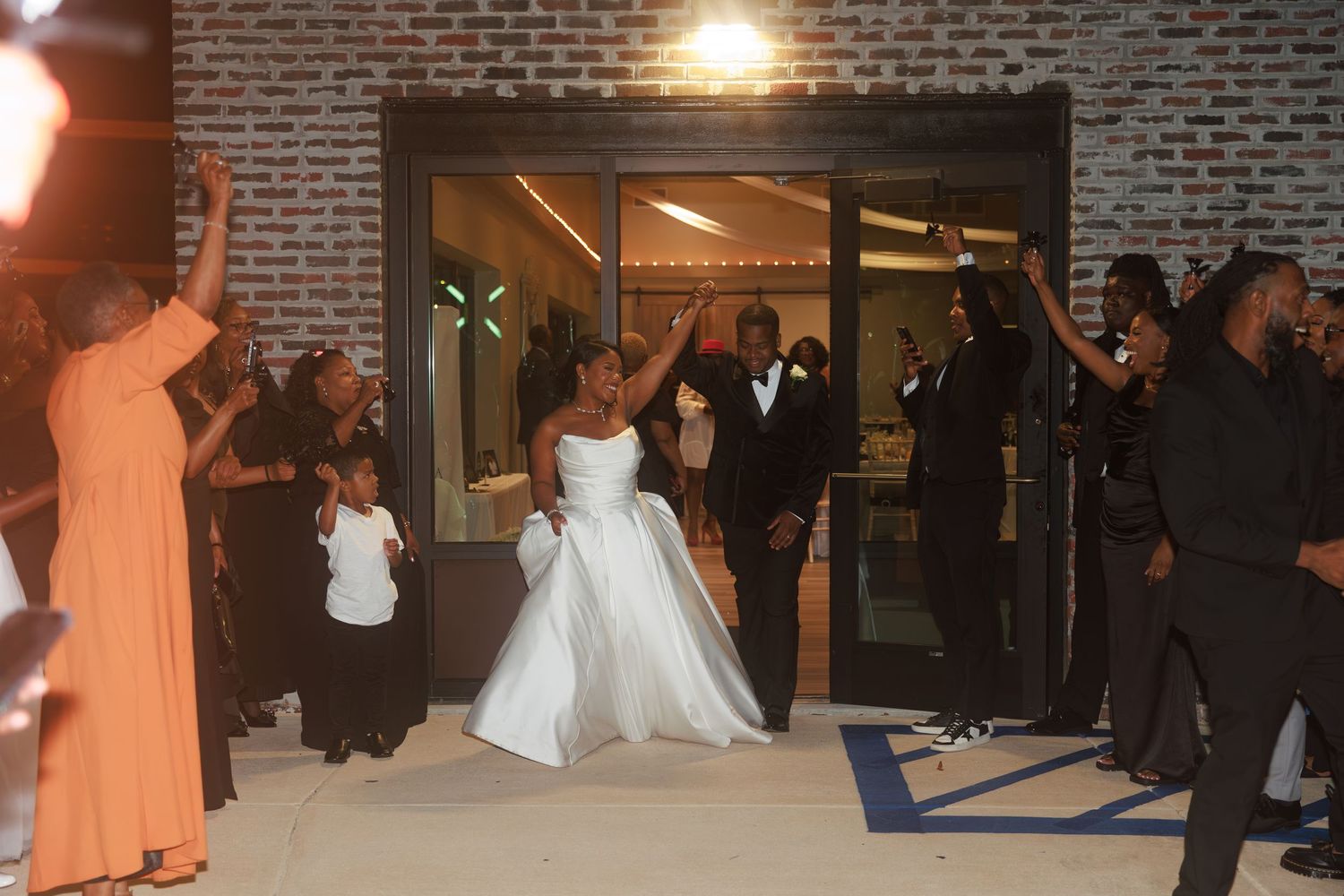 Wedding party members in orange dresses and formal attire gather outside a brick building entrance in the evening.