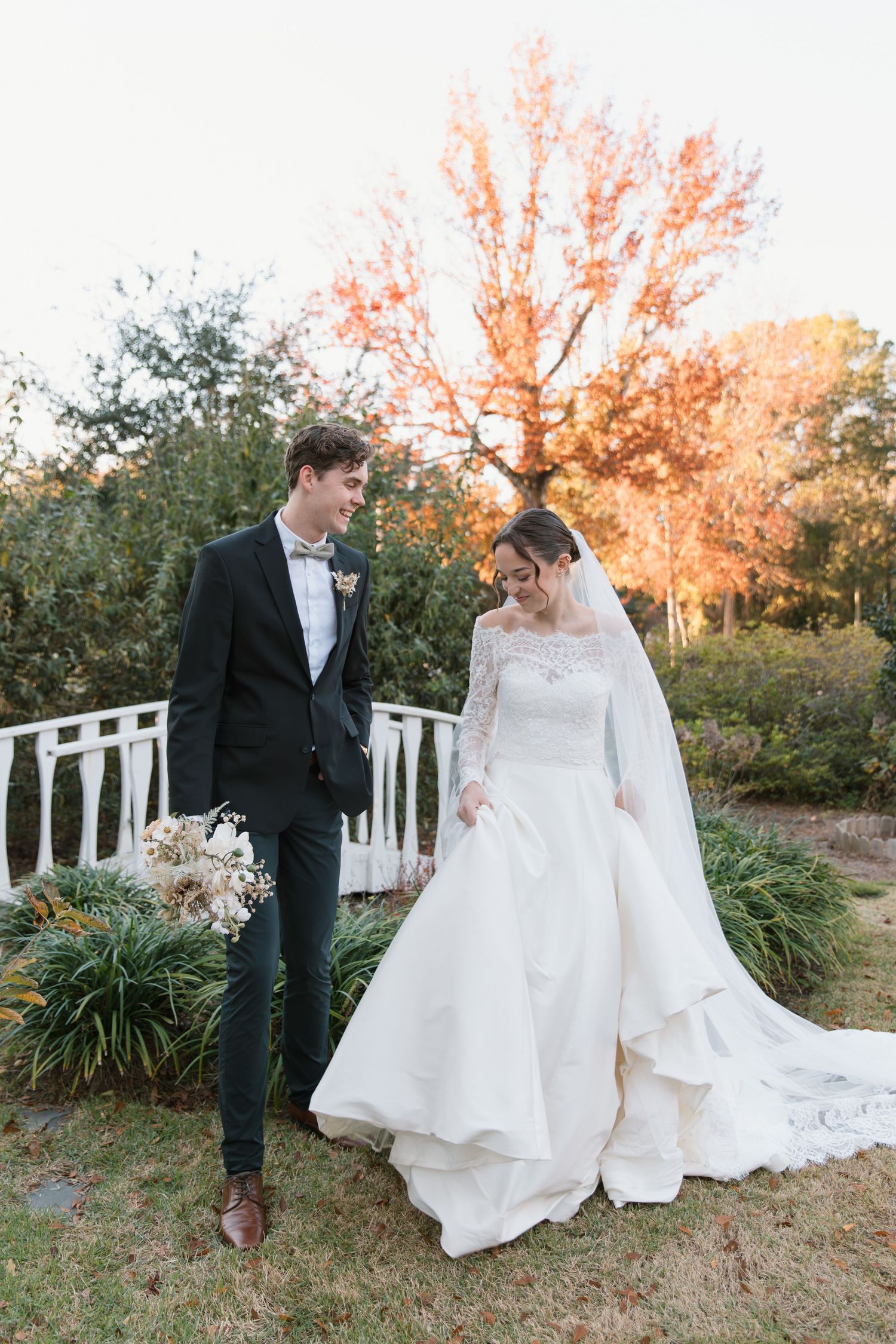 Newlywed couple shares romantic moments in garden setting with autumn trees and white fence during sunset.