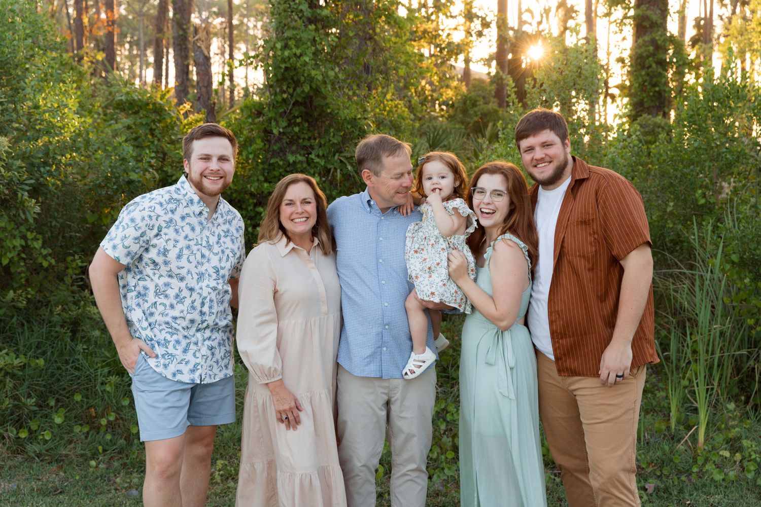 A group poses together outdoors at sunset in a wooded area wearing coordinated pastel and neutral colored outfits.