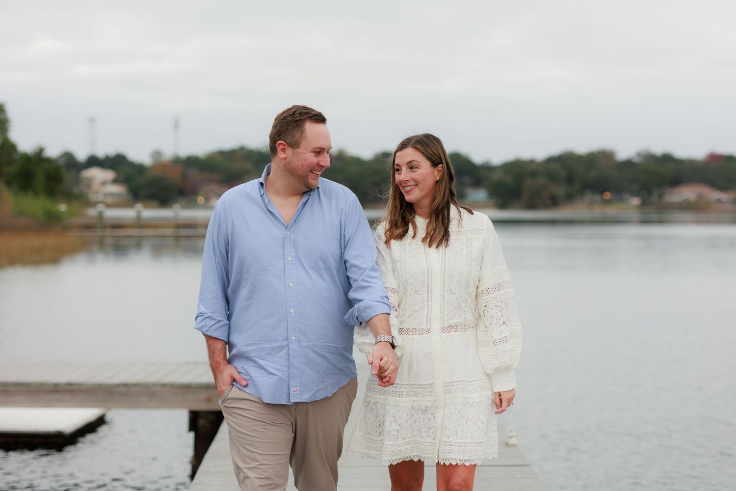 Couple walks hand in hand along scenic lakeside path during sunset engagement photoshoot.