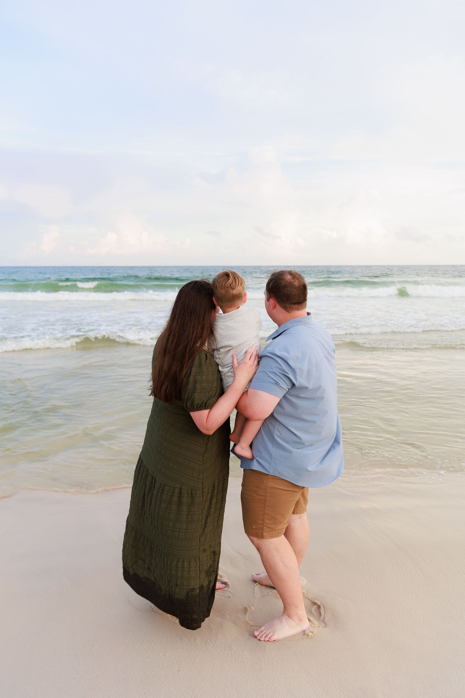 A couple stands together on a beach at sunset, looking out at the ocean waves while holding their young child.