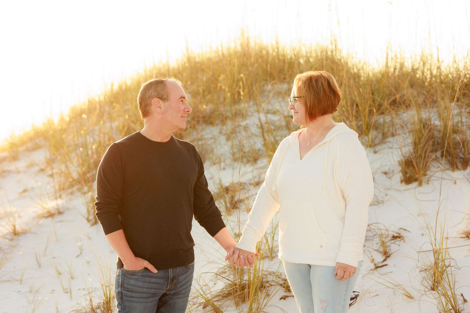 A couple holds hands while walking along a sandy beach at sunset with warm golden lighting.