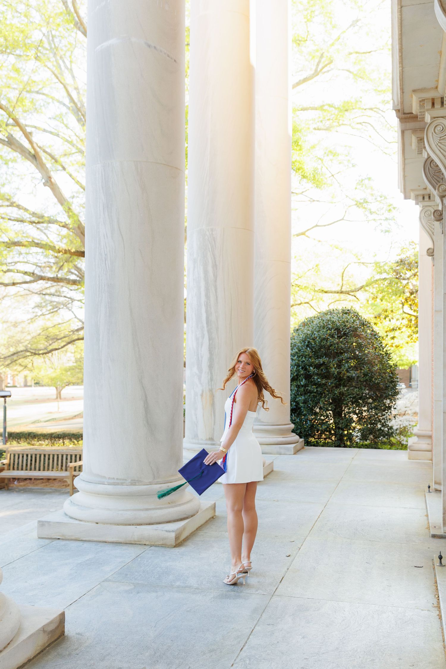 Young woman in white dress holding graduation cap walks between marble columns on a sunny college campus.