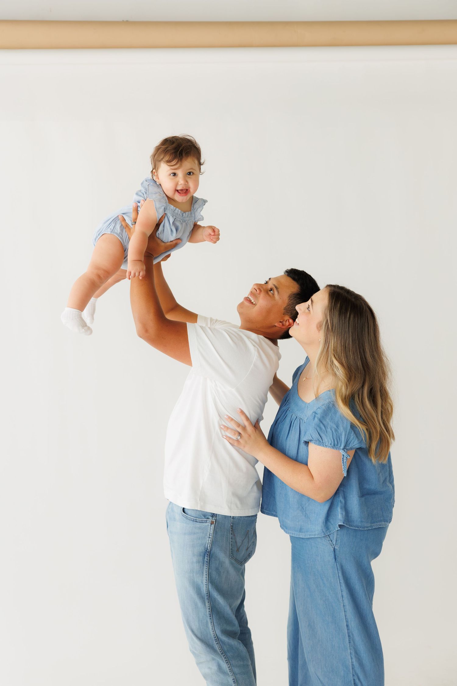 Happy parents in denim outfits lifting their smiling baby girl in the air against a white background.
