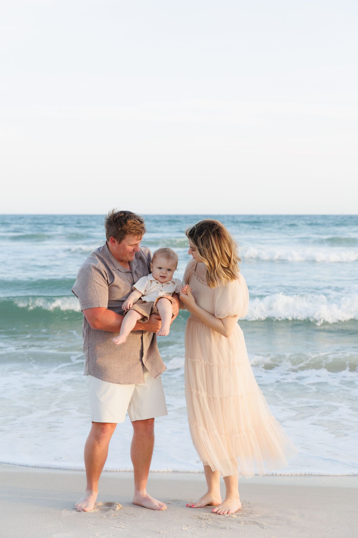 A family of three stands barefoot on a sandy beach, with a father holding a baby while the mother in a flowing dress looks on.