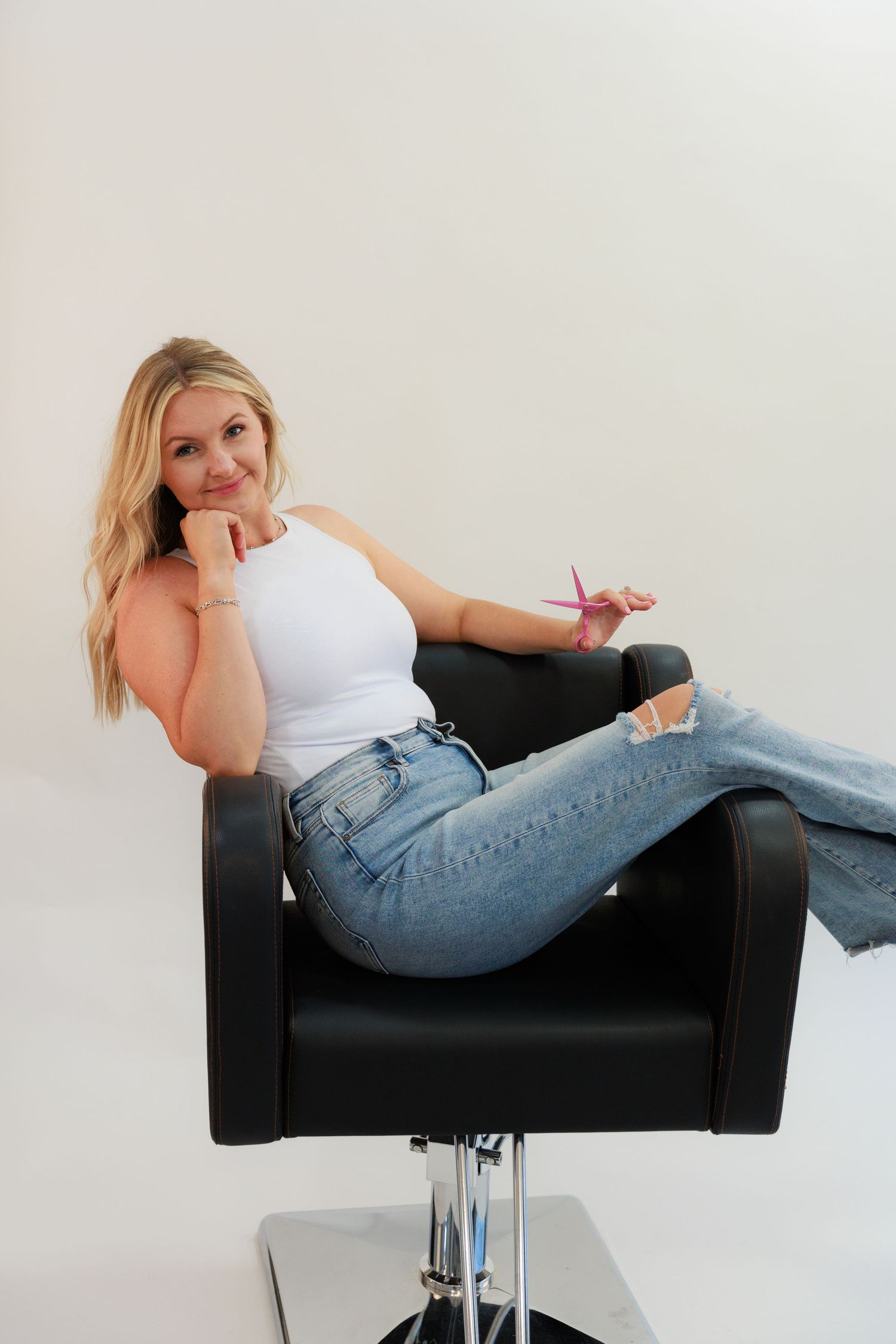 Woman in white tank top and jeans relaxing in black salon chair.