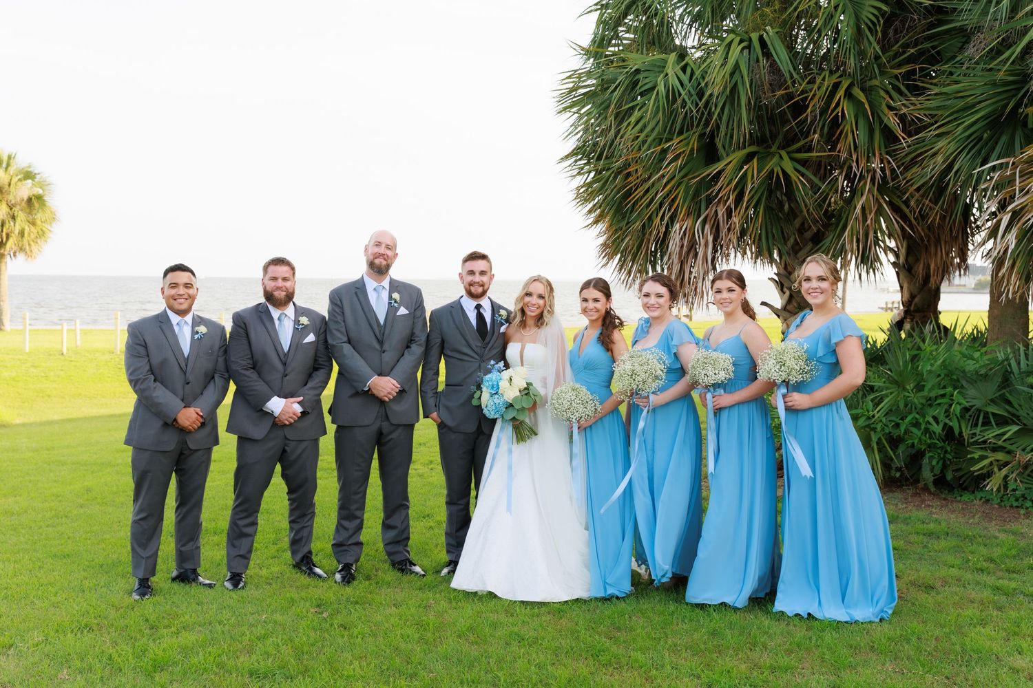 Wedding party in gray suits and light blue bridesmaid dresses poses together on grass with palm trees in background.