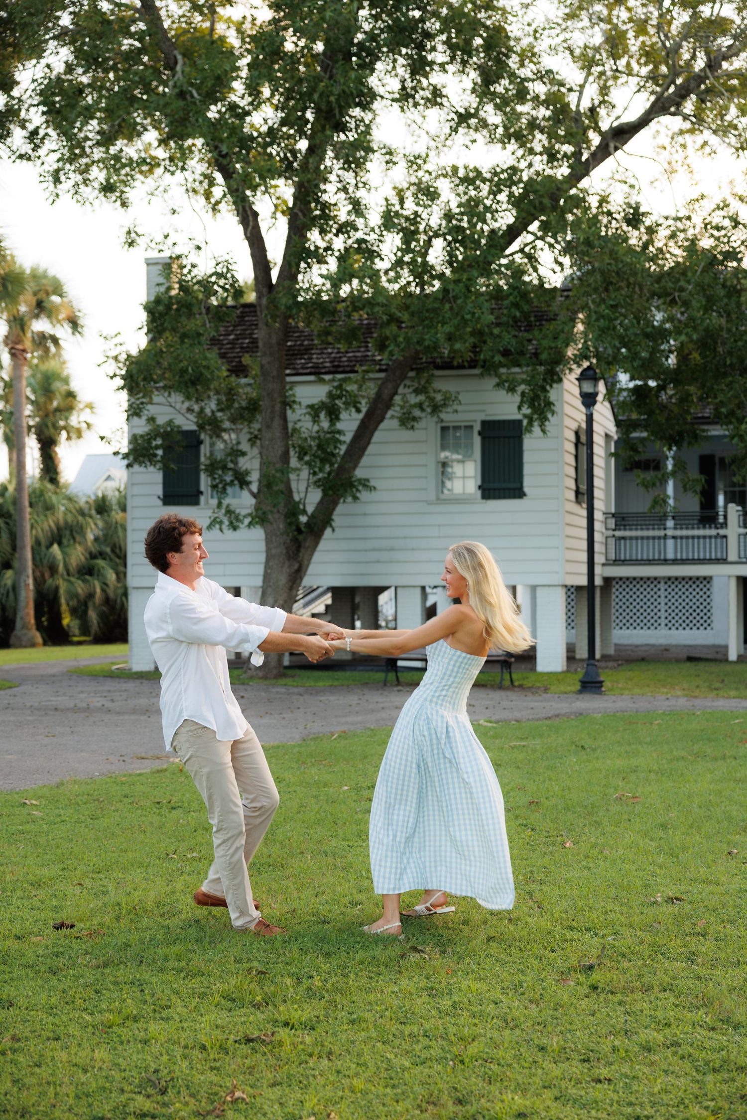 A couple spins together holding hands on a lush lawn with a white colonial building in the background.