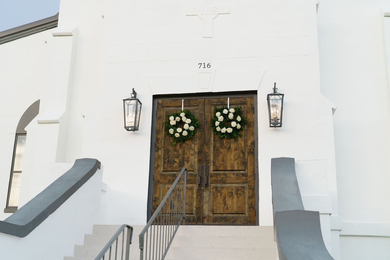 White church entrance at 716 with wooden double doors adorned with floral wreaths and lantern sconces.