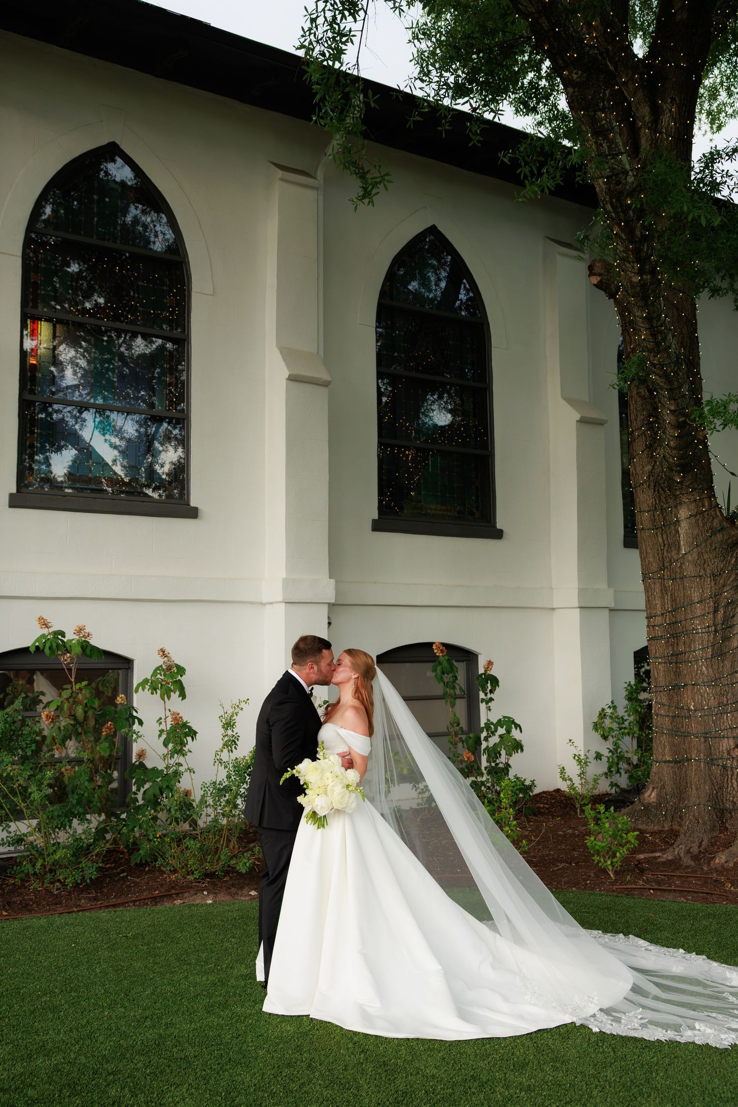 Bride and groom kissing outside white church, long veil trailing on green grass.