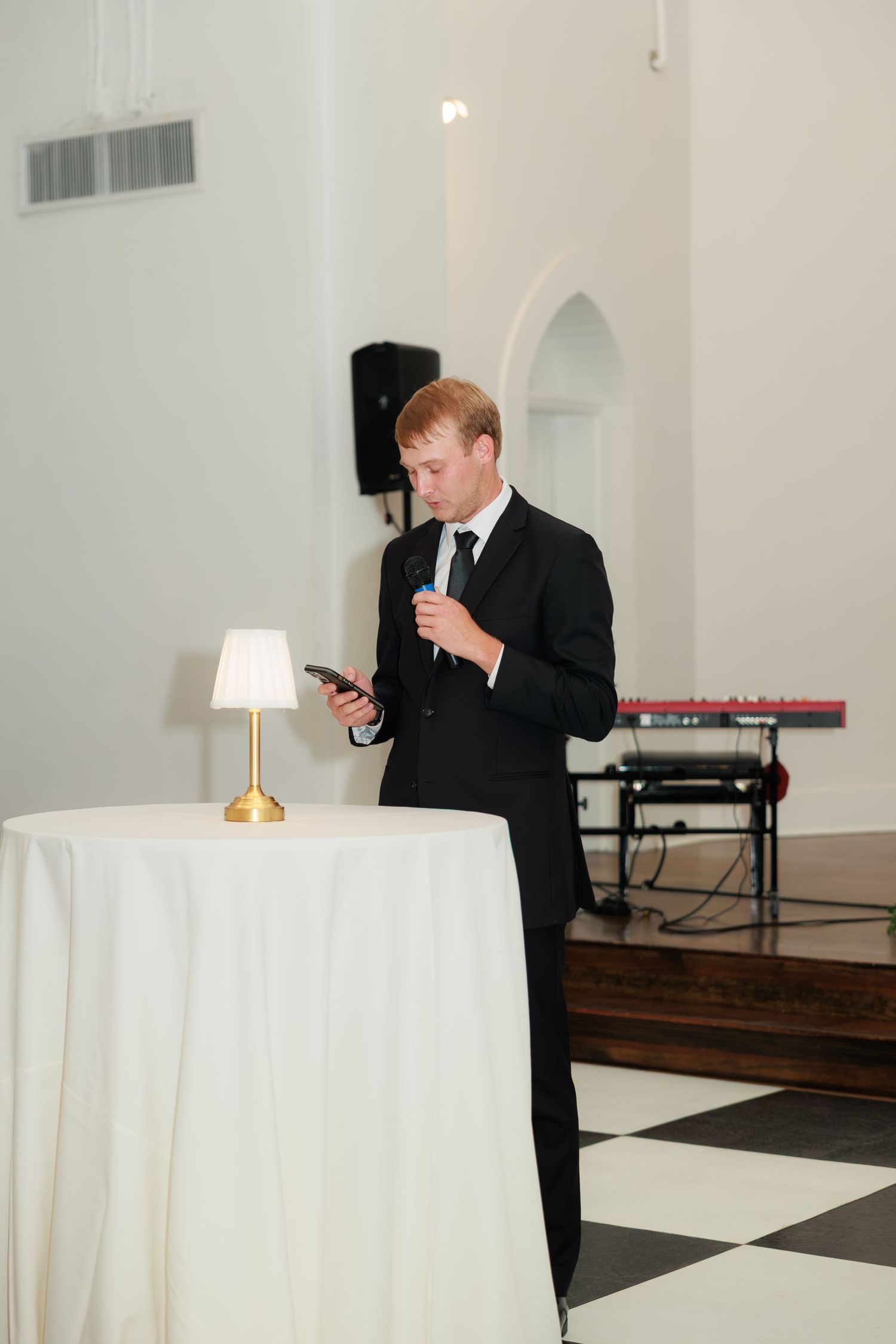 Groom in black suit reading from phone while giving speech at wedding reception.