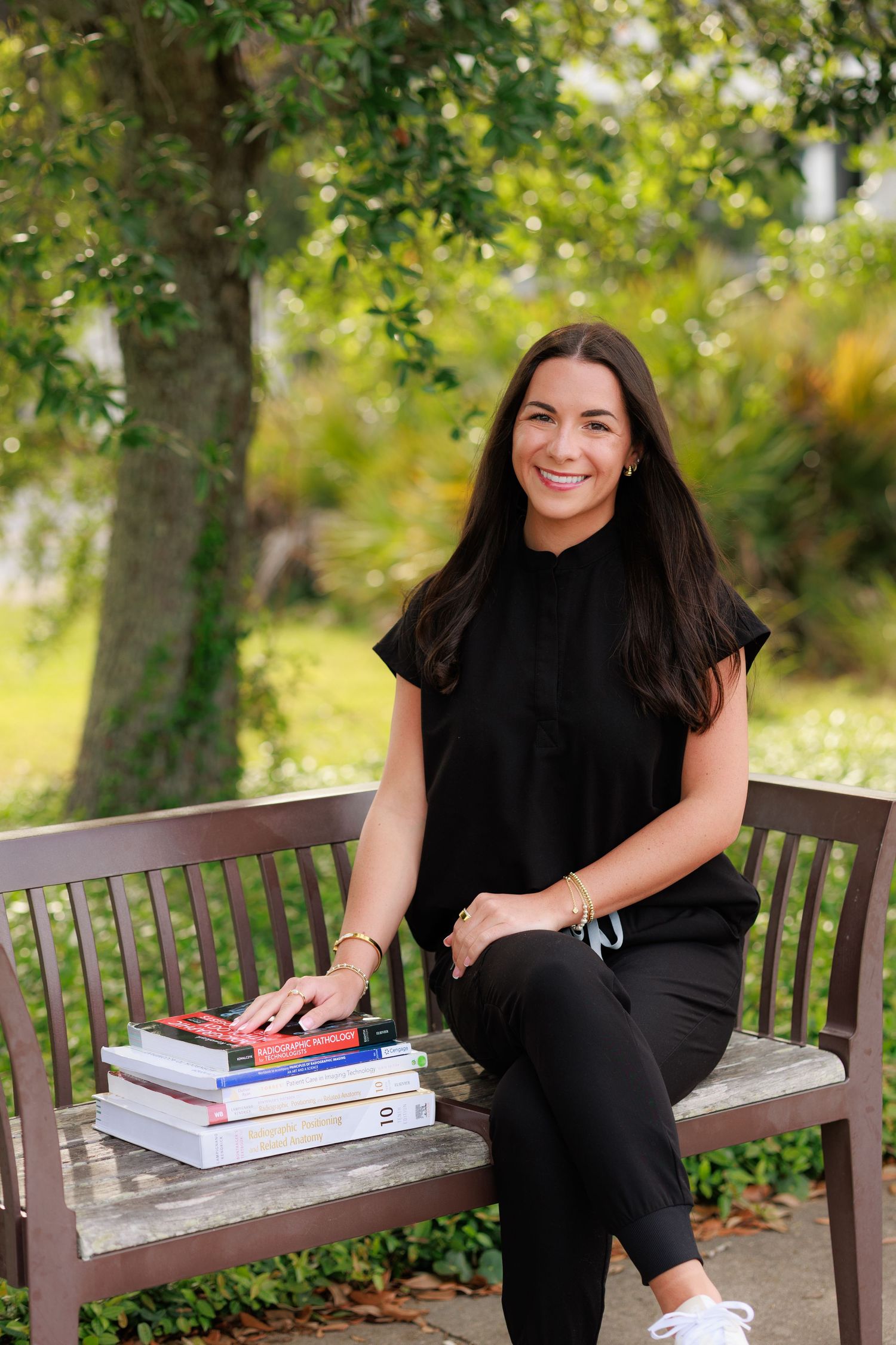 Woman in black outfit sitting on bench with stack of books outdoors, smiling in a lush green park setting.