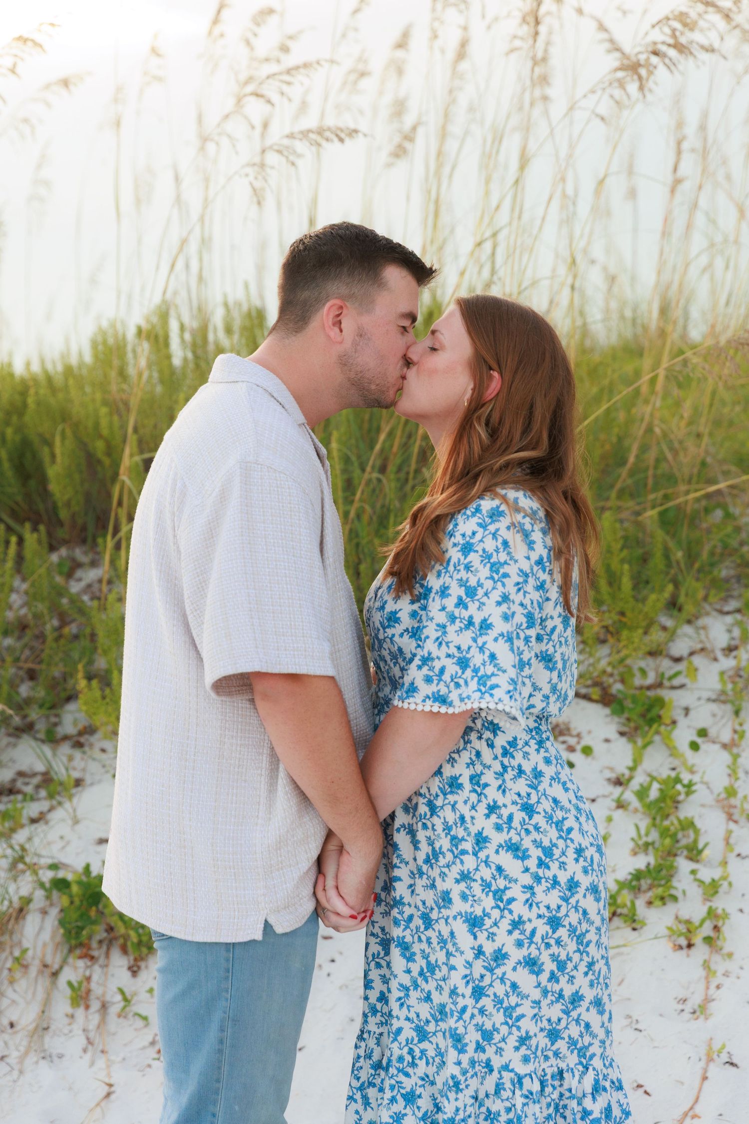 A couple shares a tender kiss while holding hands on a white sand beach surrounded by sea oats.