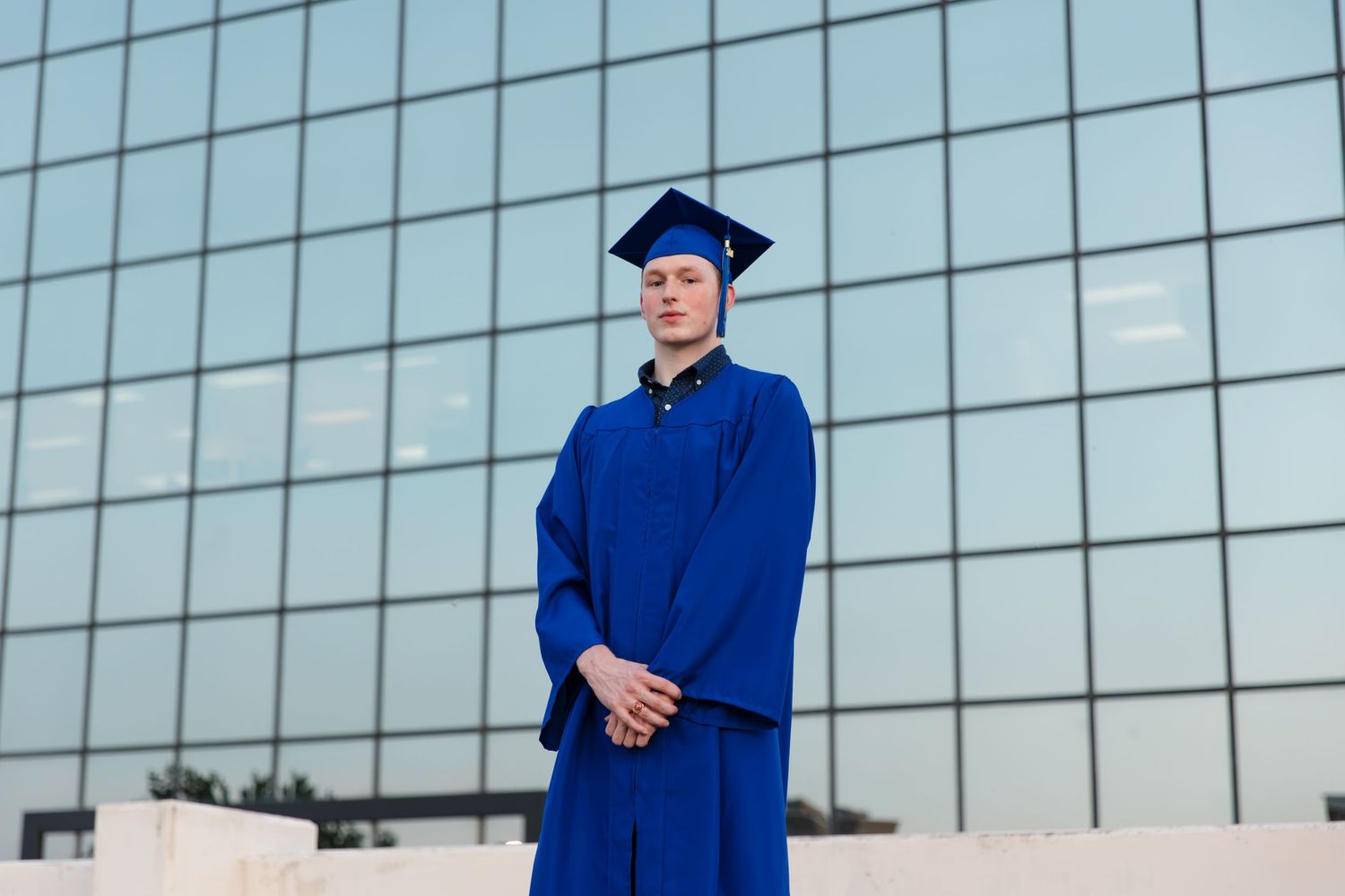 Young graduate in blue cap and gown stands confidently before a modern glass building.