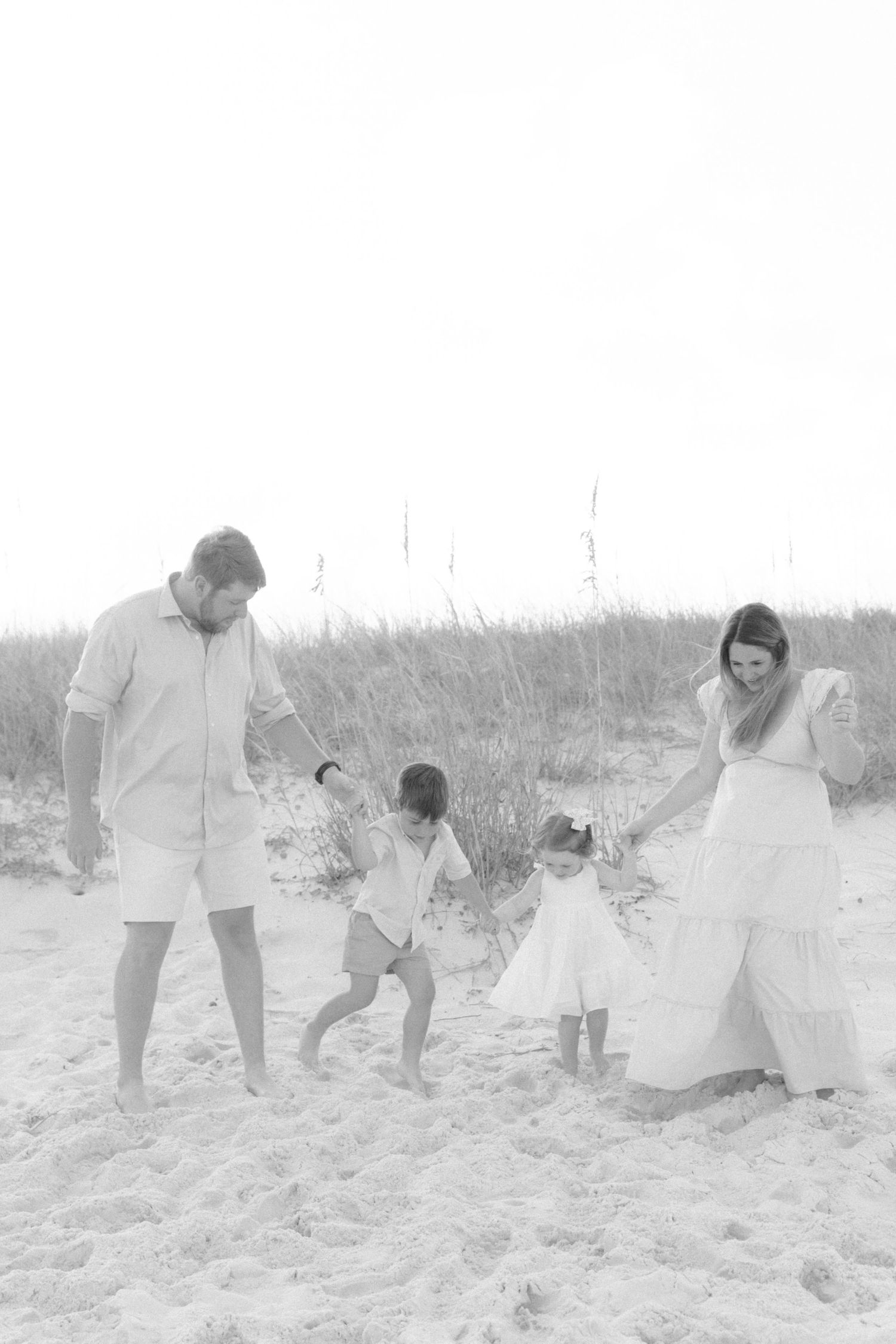Black and white photo of a family of four playing on a sandy beach near sea oats, parents holding hands with two young girls.