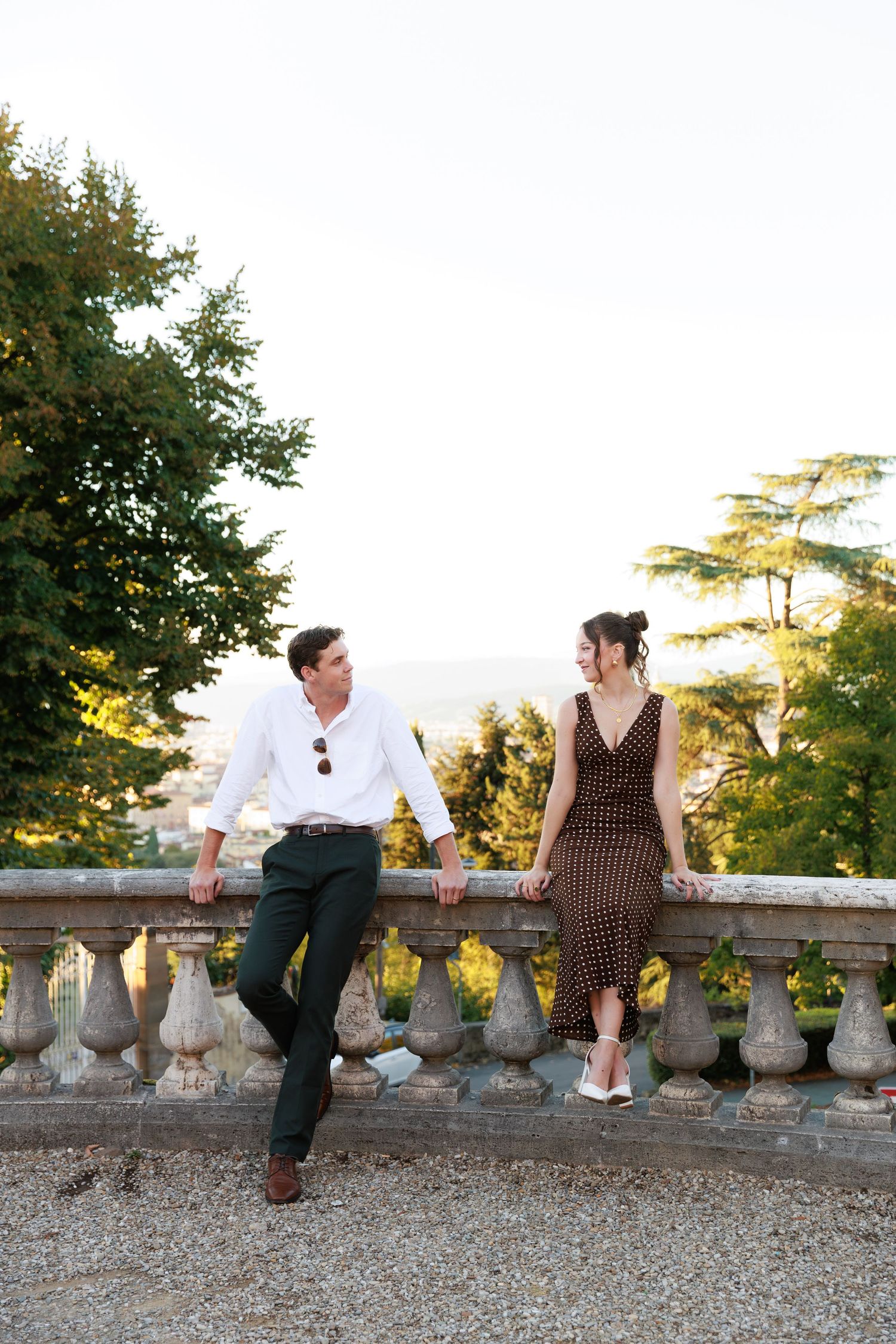 Couple in elegant attire leaning against stone balustrade in lush garden, sharing a romantic glance at golden hour.