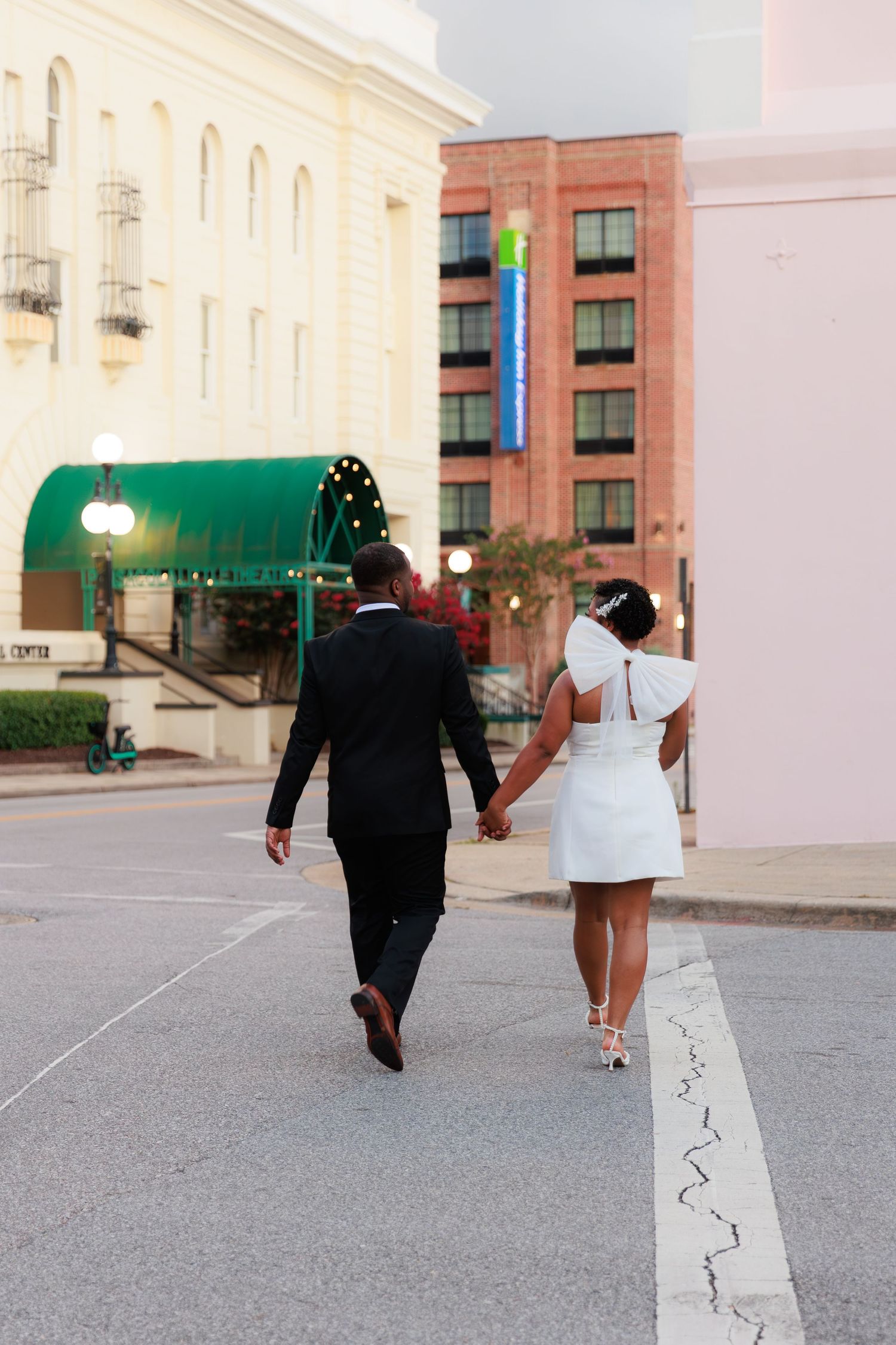A couple in wedding attire walks hand-in-hand down a city street, the bride in a white mini dress and groom in a black suit.